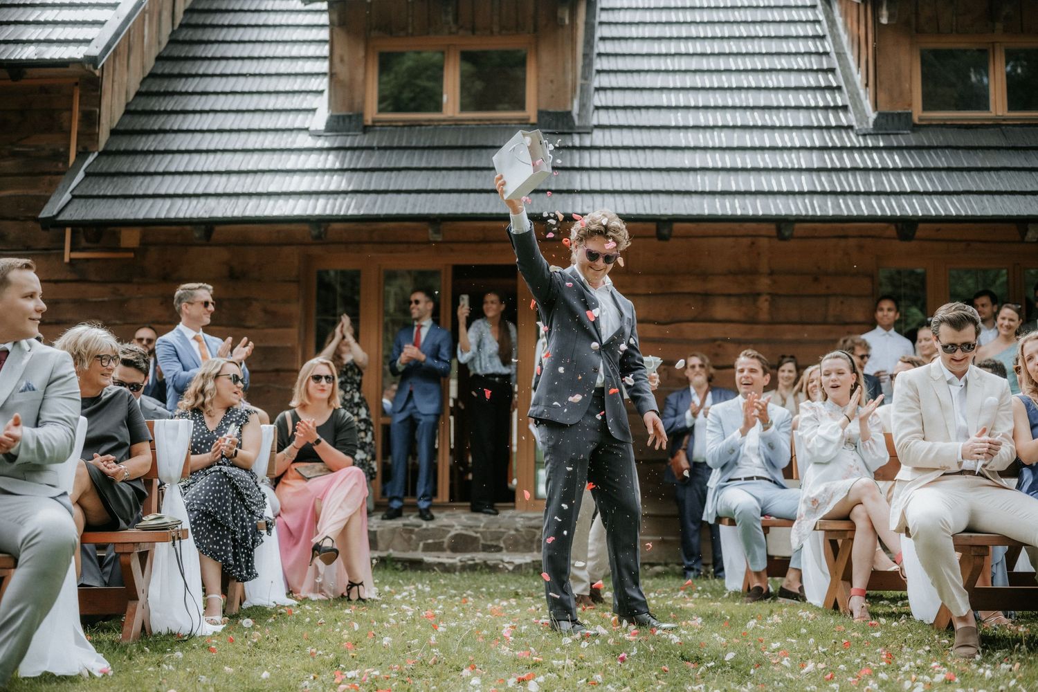 Groom walks down aisle tossing flower petals at outdoor rustic wooden lodge wedding ceremony with guests applauding.