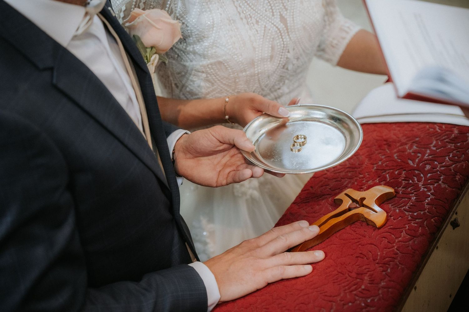 Bride and groom exchanging wedding rings on silver plate during religious ceremony with wooden cross.
