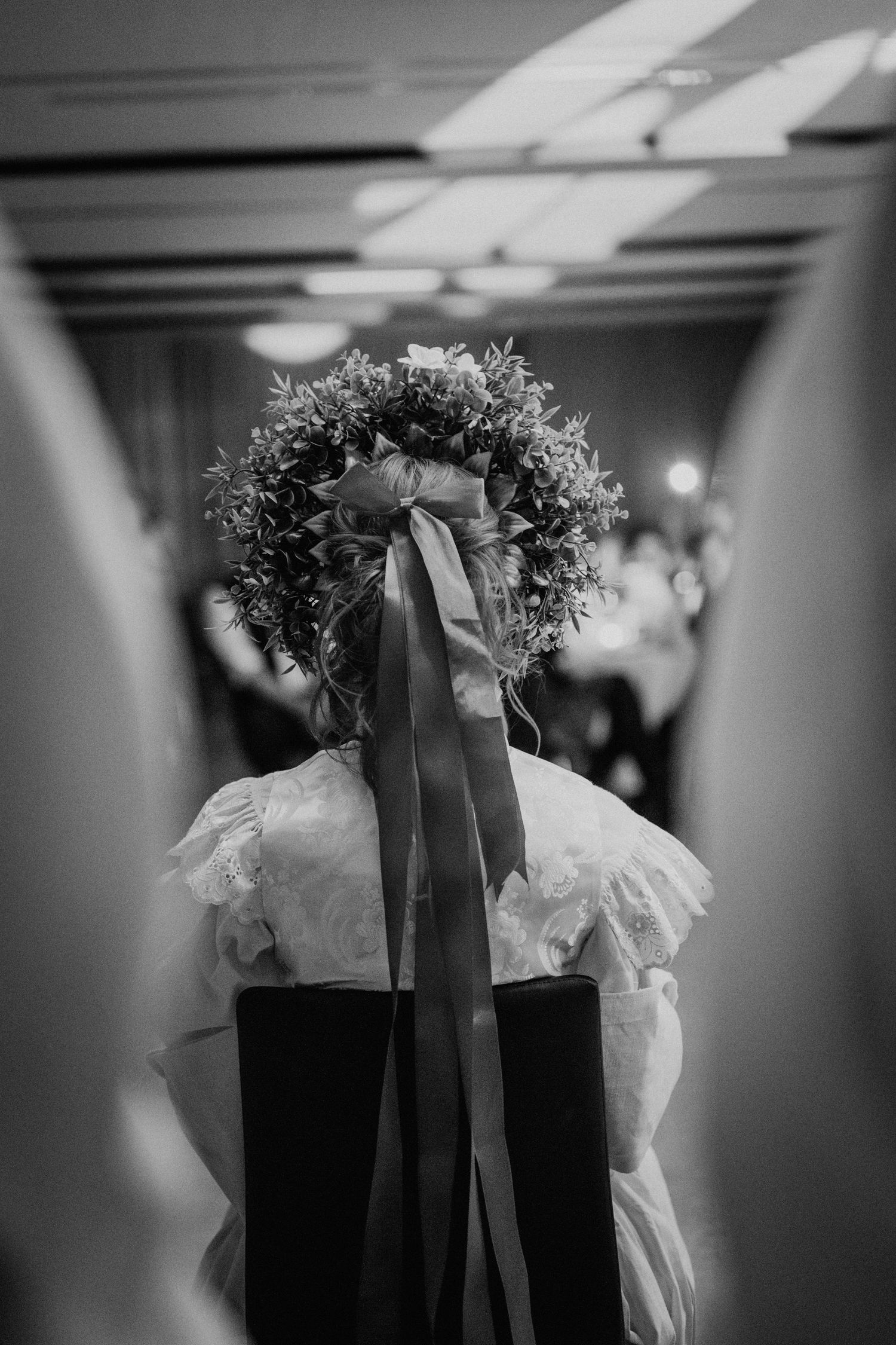 Black and white photo of a child wearing a floral crown with long ribbons, viewed from behind at a formal event.