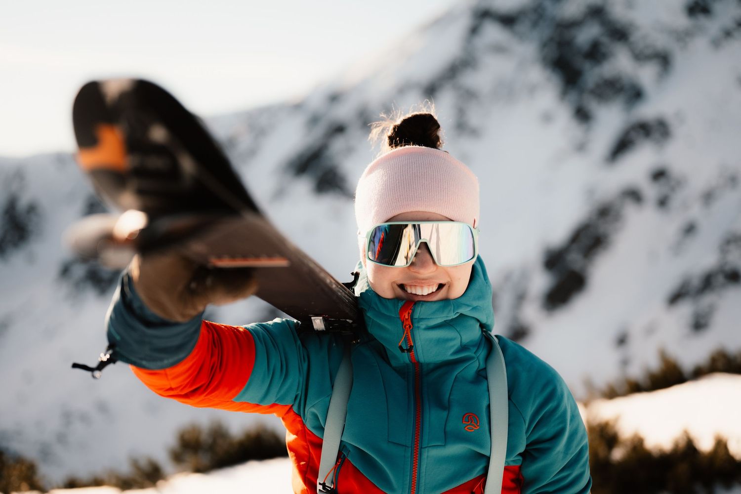 Female skier in teal jacket holding ski overhead with snowy mountain backdrop on sunny winter day.