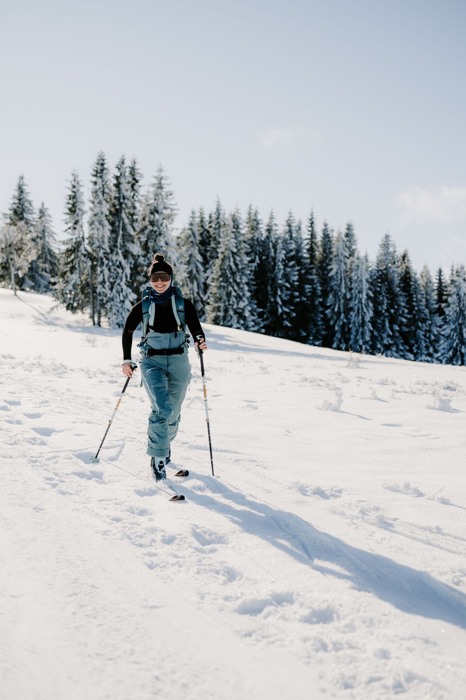Female backcountry skier ascending a snowy slope with snow-laden pine trees in the background.