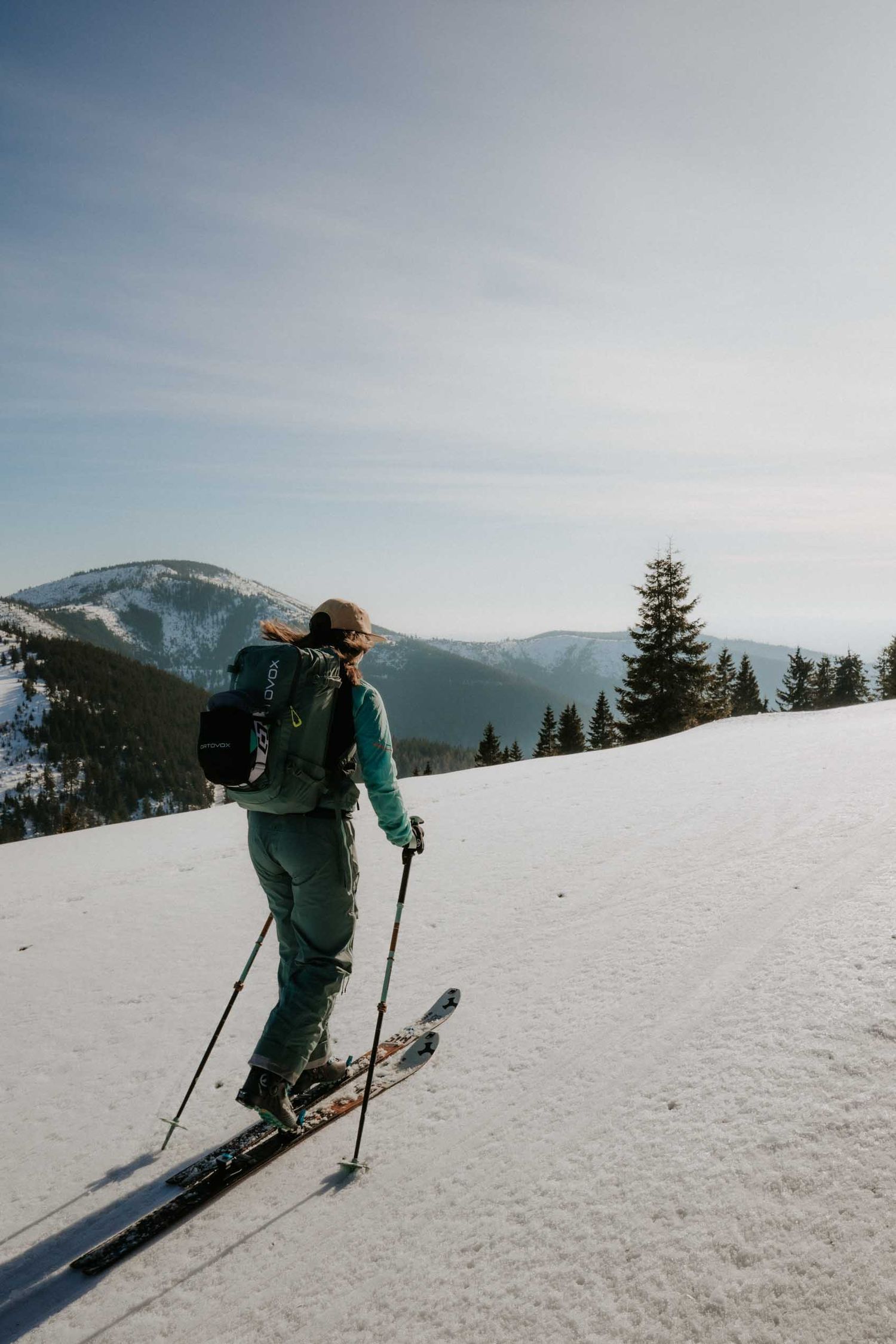 Female skier in teal outfit ski touring on snowy mountain slope with pine trees and peaks.