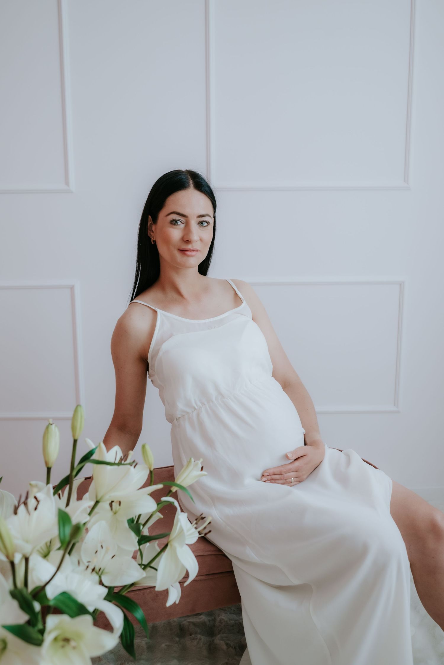 Smiling pregnant woman in white dress sitting on ottoman with white lilies in foreground.