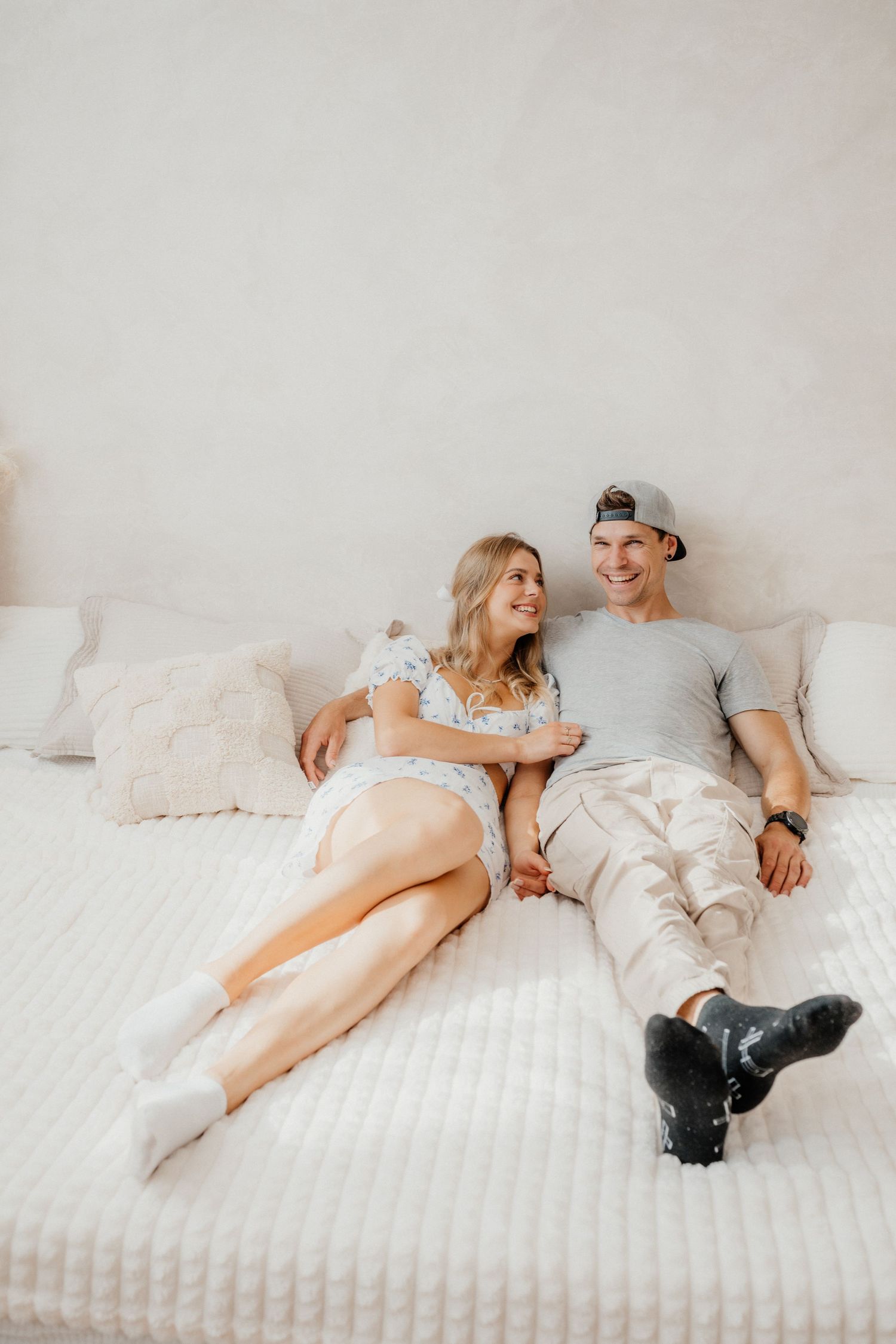 A relaxed couple sits together on a white bed, smiling at each other in a bright minimalist bedroom setting.