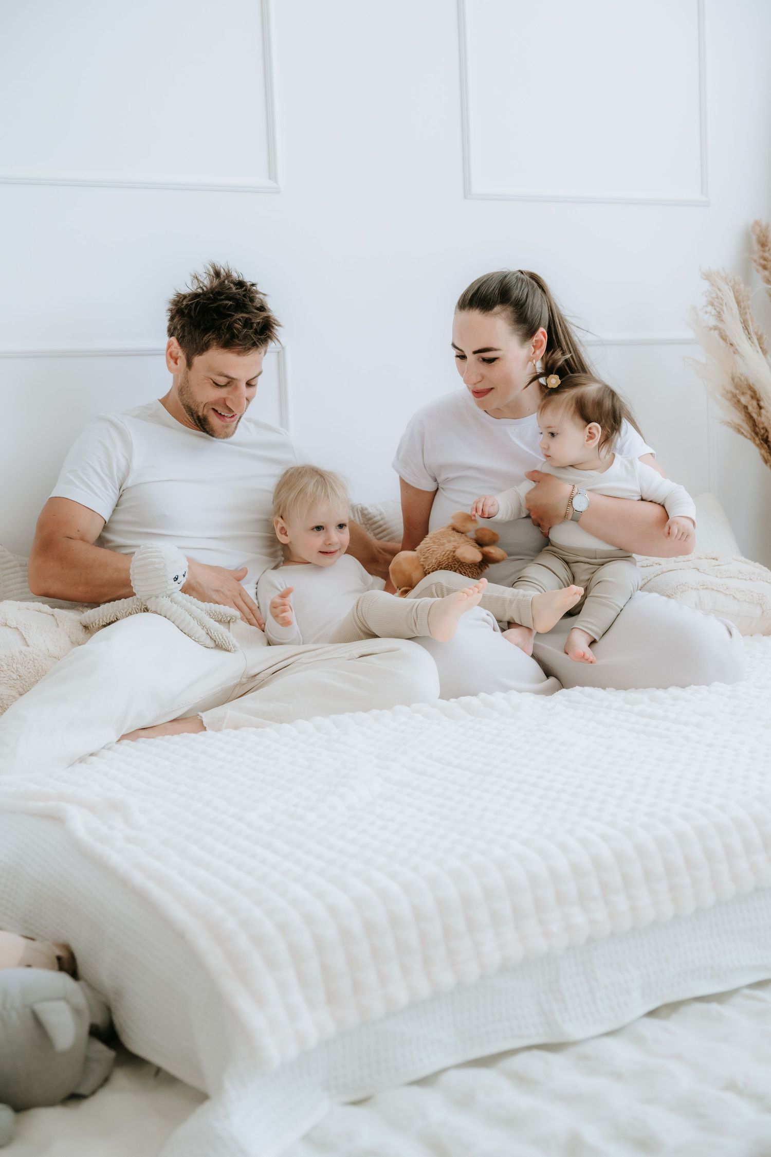 Happy family of four in white outfits sitting on bed with teddy bear in bright room.