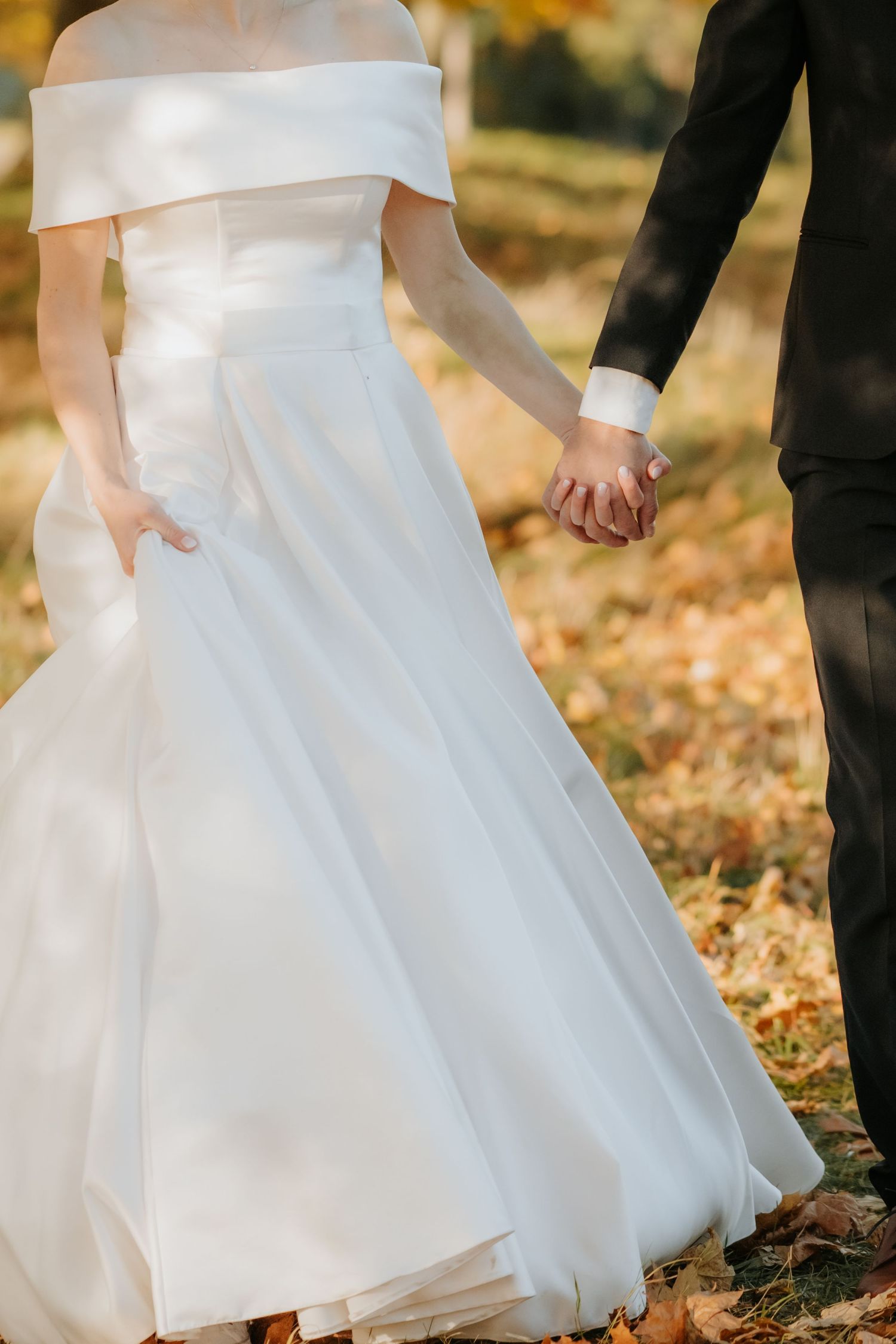 Bride in white off-shoulder gown holds groom's hand in black suit among autumn leaves.