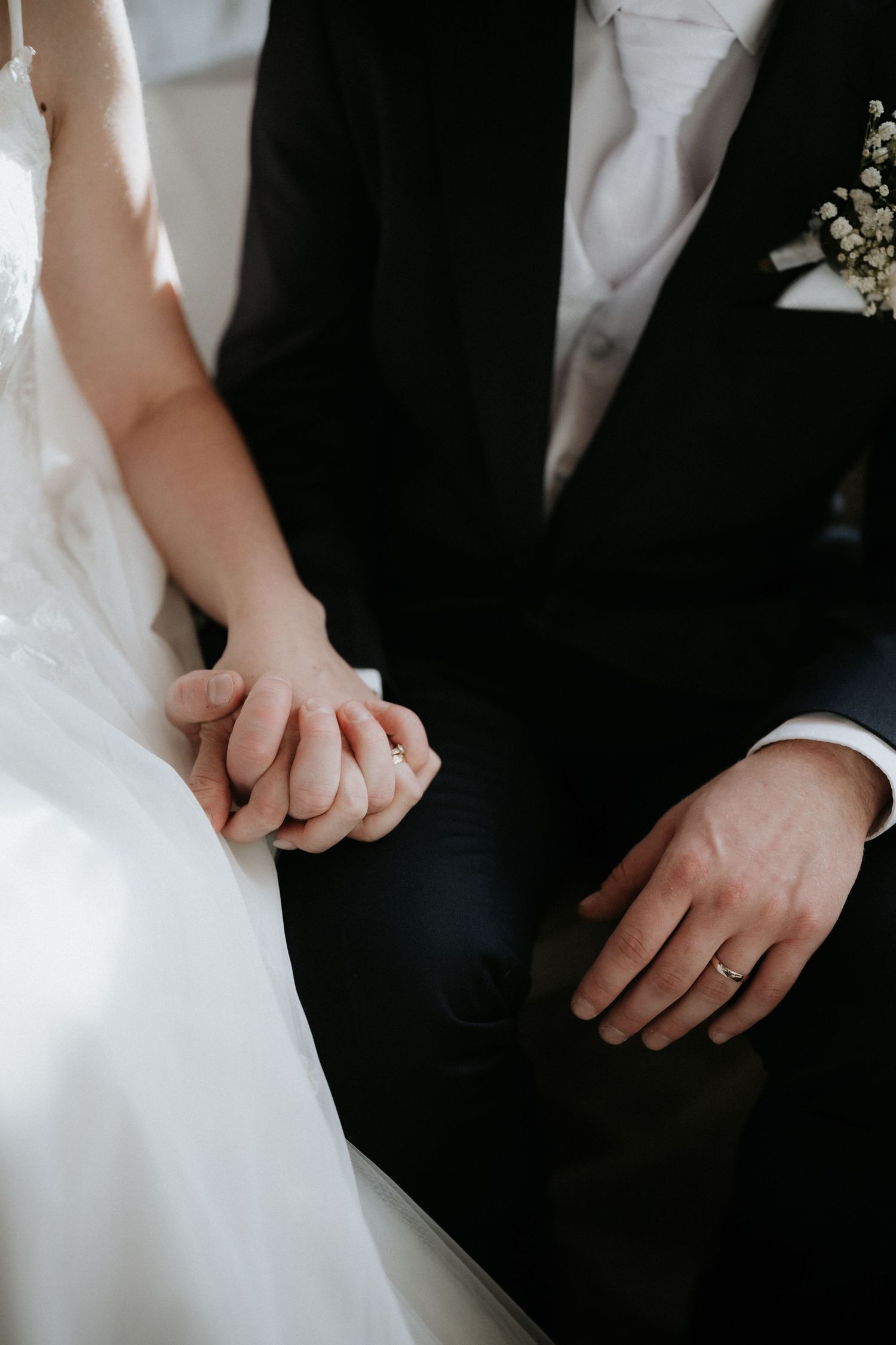Bride and groom holding hands, showing wedding rings, bride in white dress, groom in black suit with floral boutonniere.