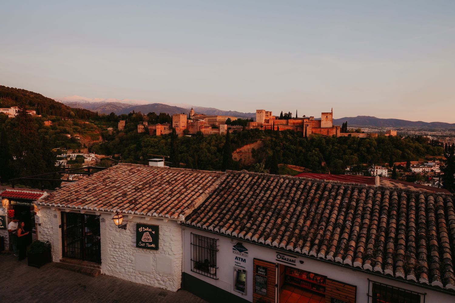 Sunset view of the Alhambra palace and Granada cityscape with terracotta rooftops and mountains in background.