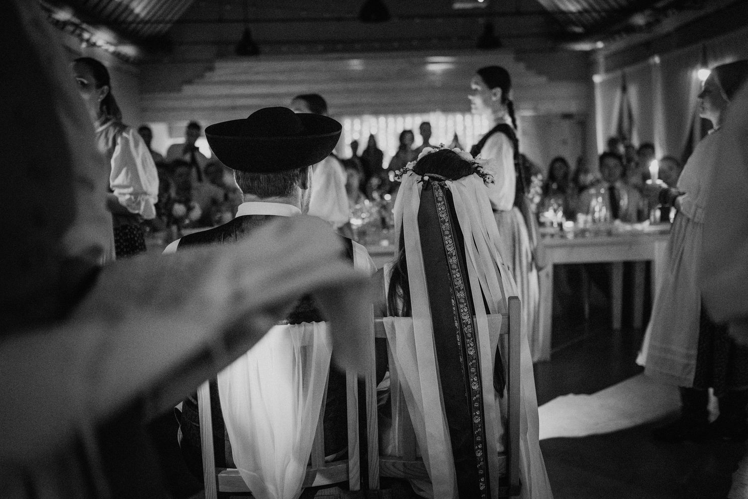 Black and white photo of guests in traditional folk costumes at a wedding reception ceremony indoors.