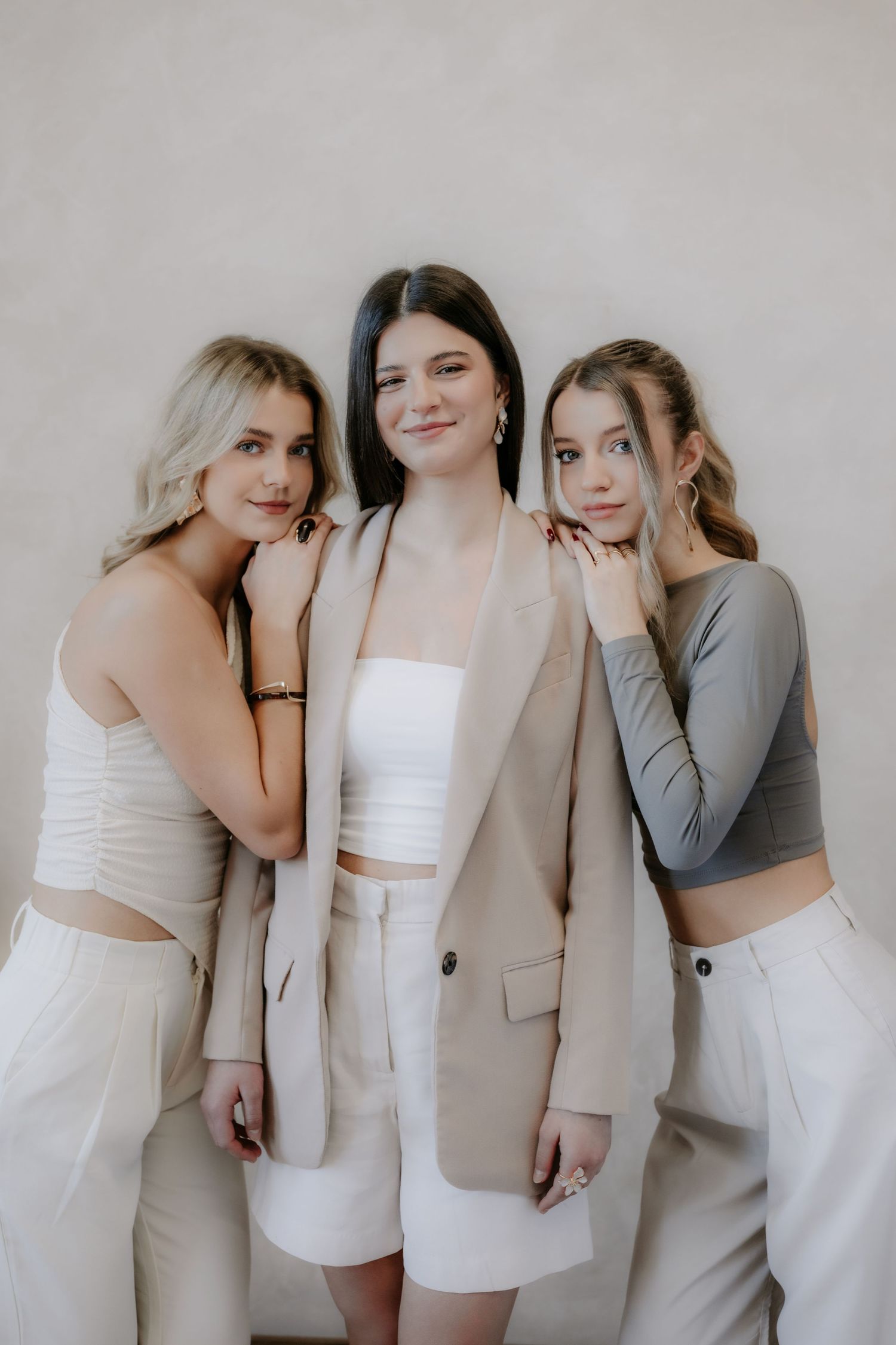Three fashionable young women in elegant neutral outfits posing together against a beige textured background.