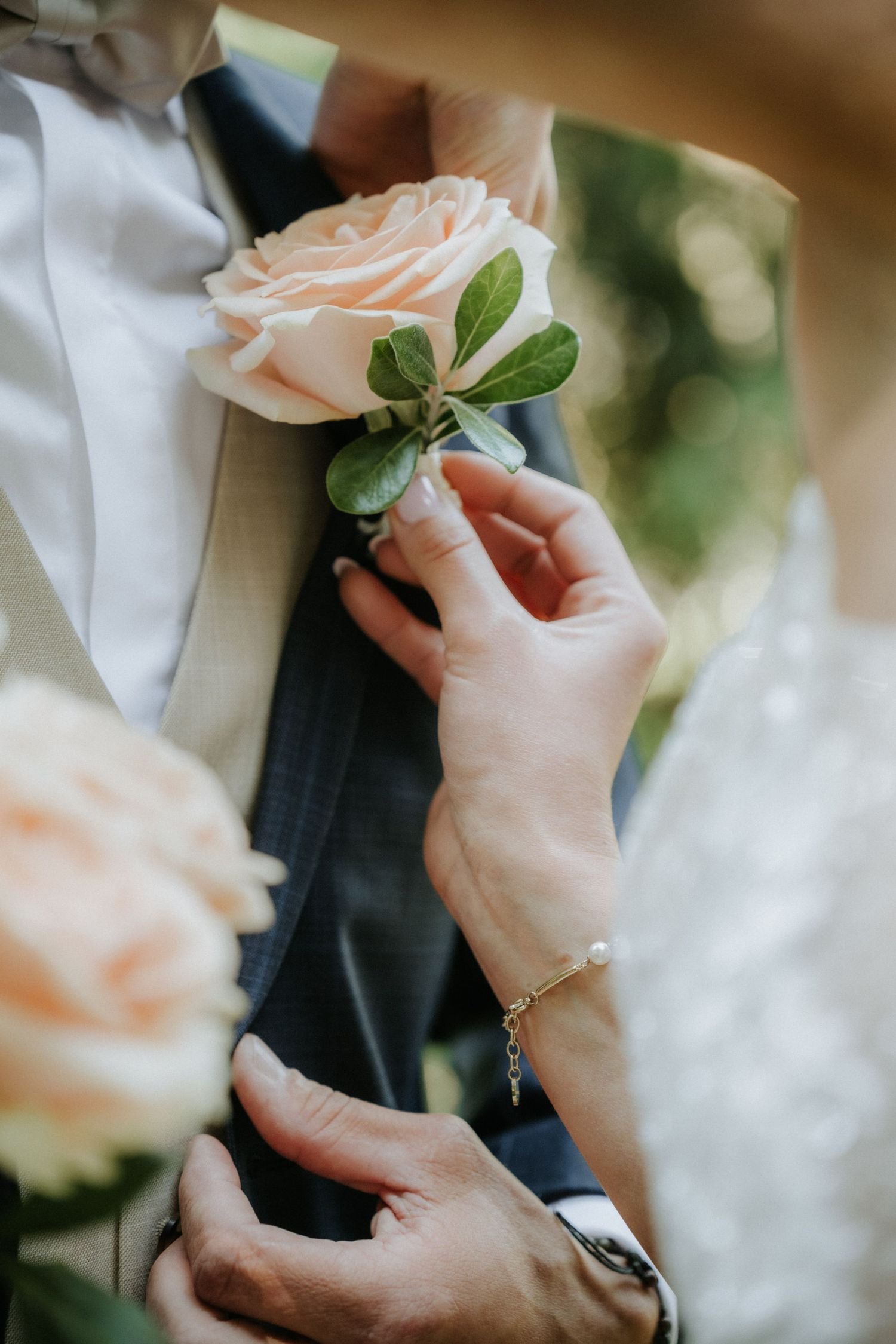 Bride pinning a peach rose boutonniere on groom's navy suit lapel at wedding ceremony.
