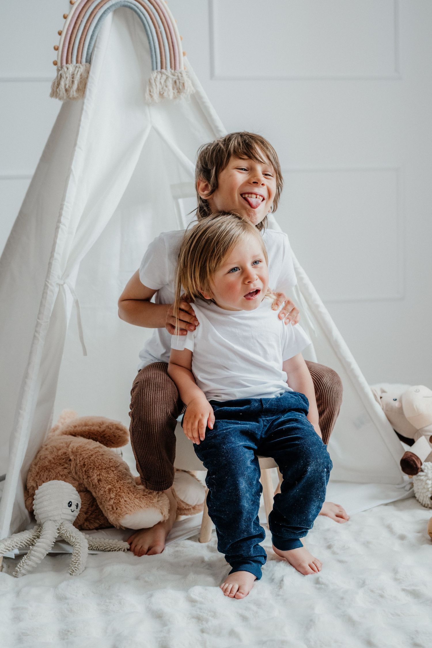 Two young brothers laugh and play together in front of a white teepee tent surrounded by stuffed toys.