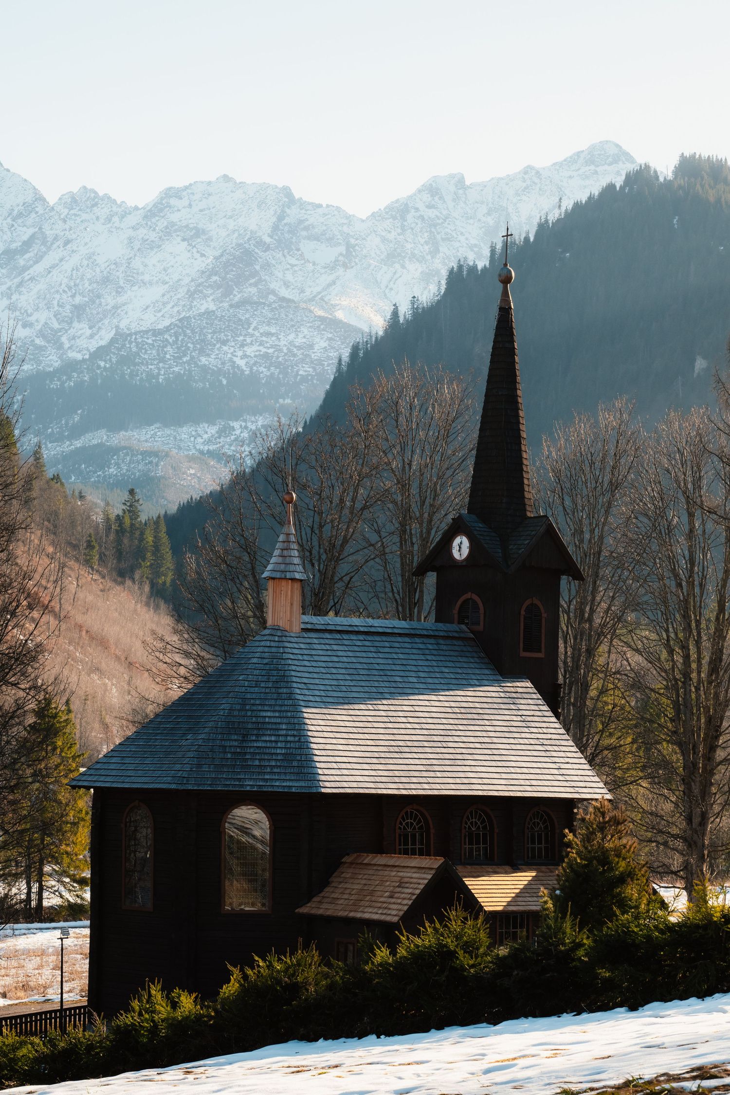 Traditional dark wooden chapel with steeple set against snow-dusted mountain peaks in winter.