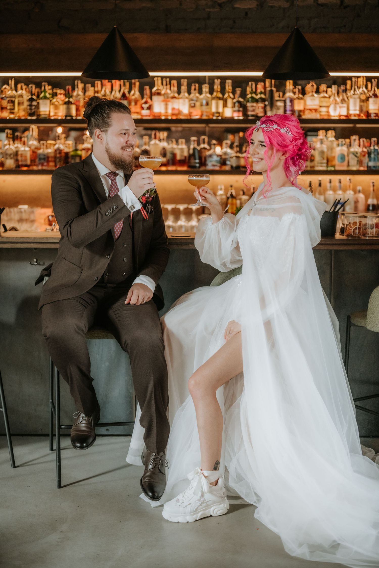 Bride with pink hair and groom in brown suit toasting cocktails at a bar on their wedding day.