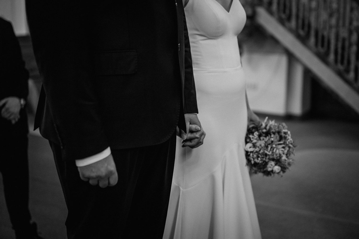 Black and white photo of a bride and groom holding hands, bride holding a floral bouquet in an elegant wedding gown.
