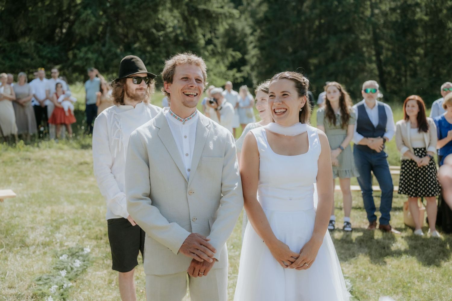 Smiling bride and groom stand outdoors at a sunny garden wedding ceremony with guests gathered in the background.