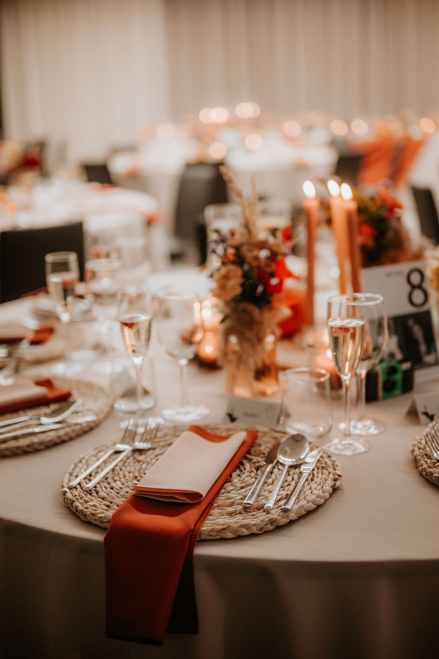 Elegant wedding reception table setting with woven chargers, terracotta napkins, crystal glasses, and floral centerpieces.