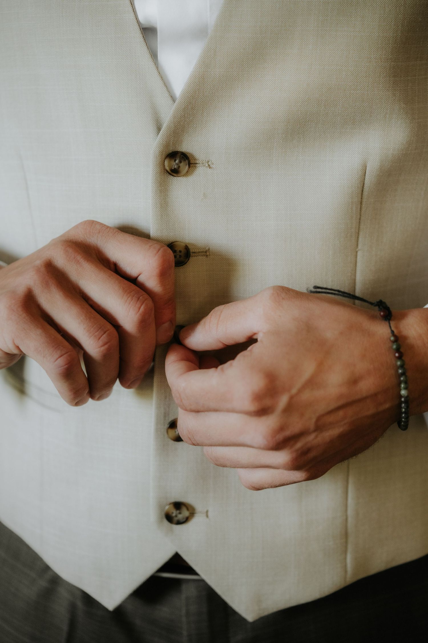 Man buttoning a beige suit vest, wearing a beaded bracelet, close-up detail shot for formal wear.