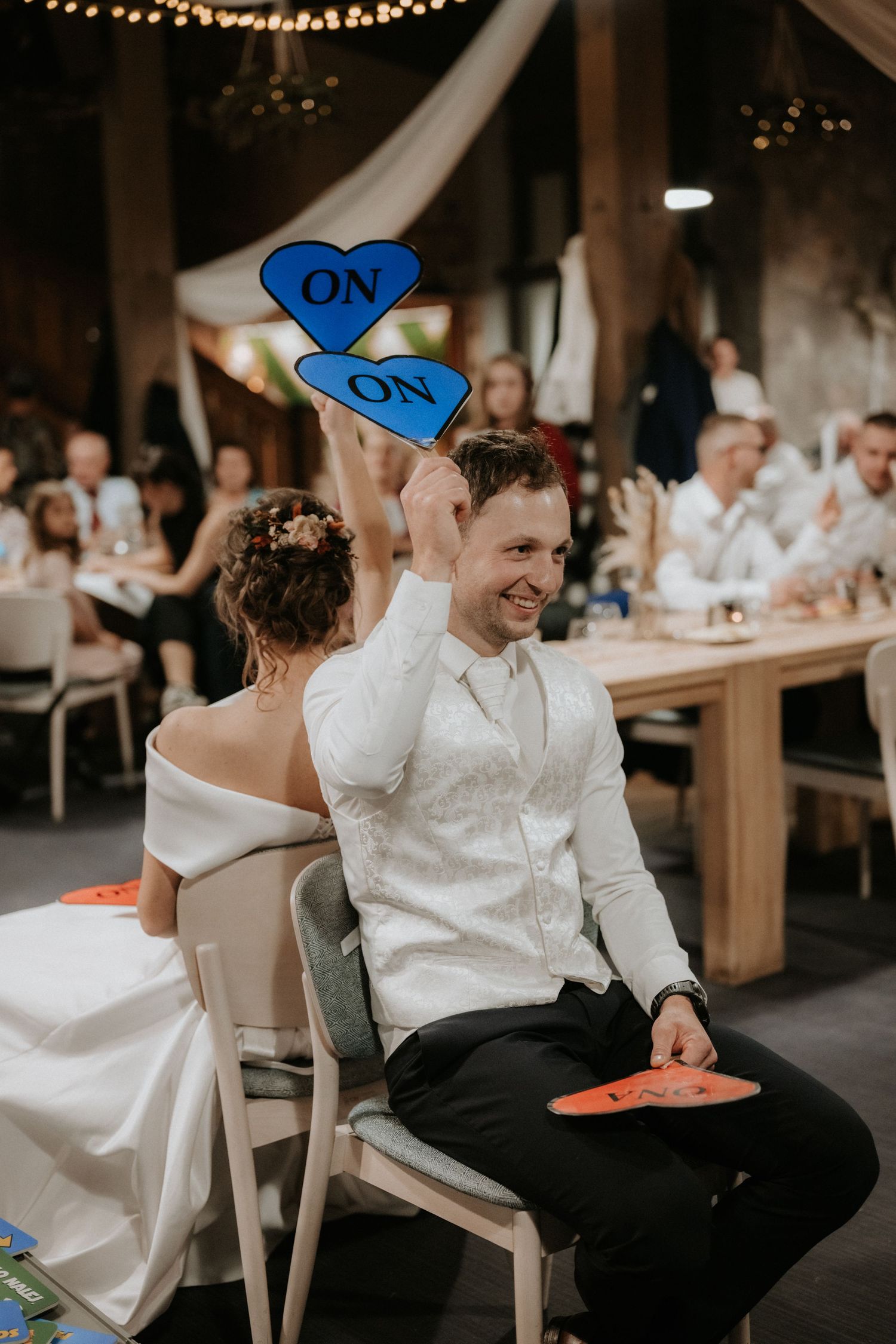 Smiling groom holds blue heart-shaped ON signs at wedding reception while bride sits beside him in elegant venue.