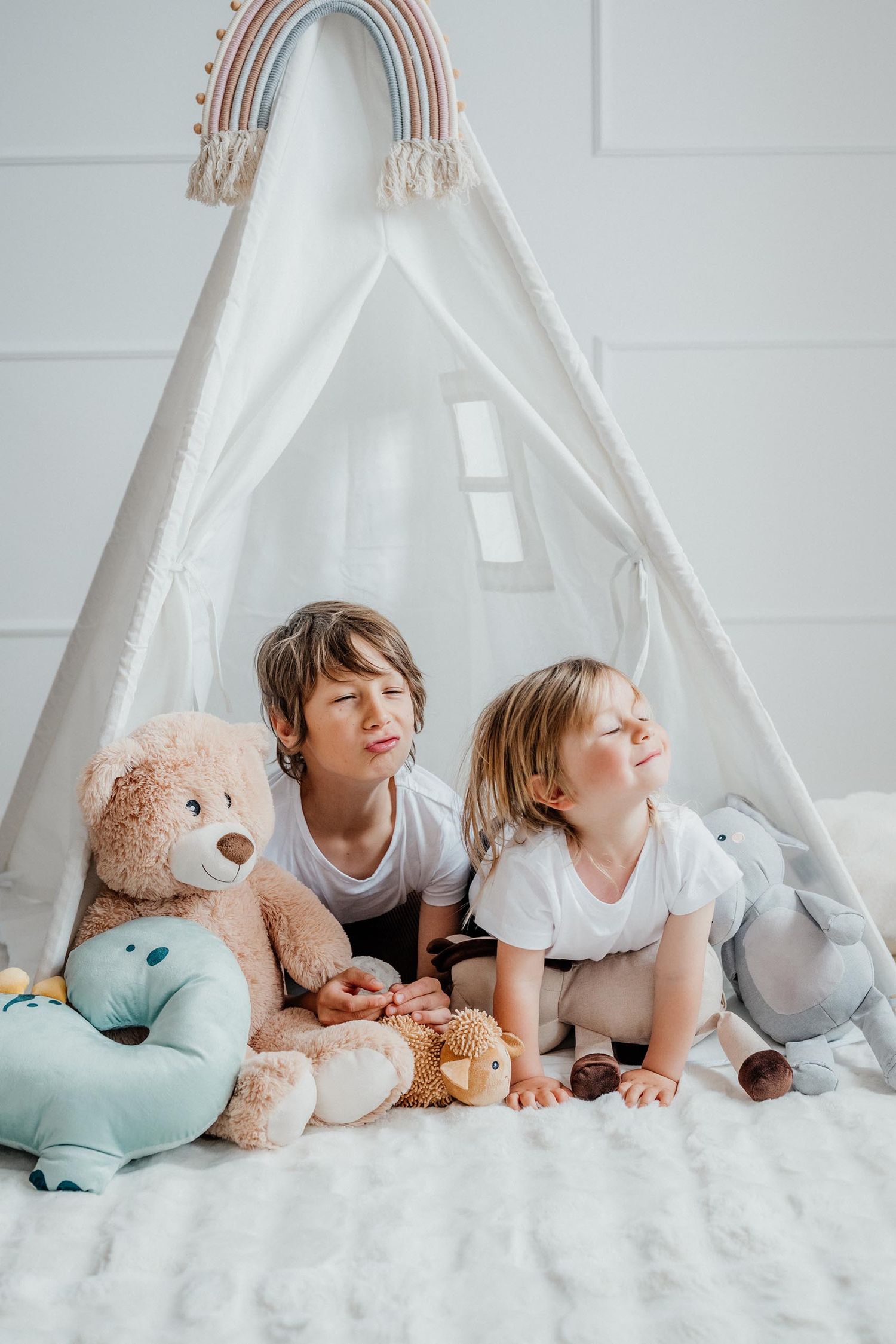 Two children sitting in a white play tent with stuffed animals and toys.