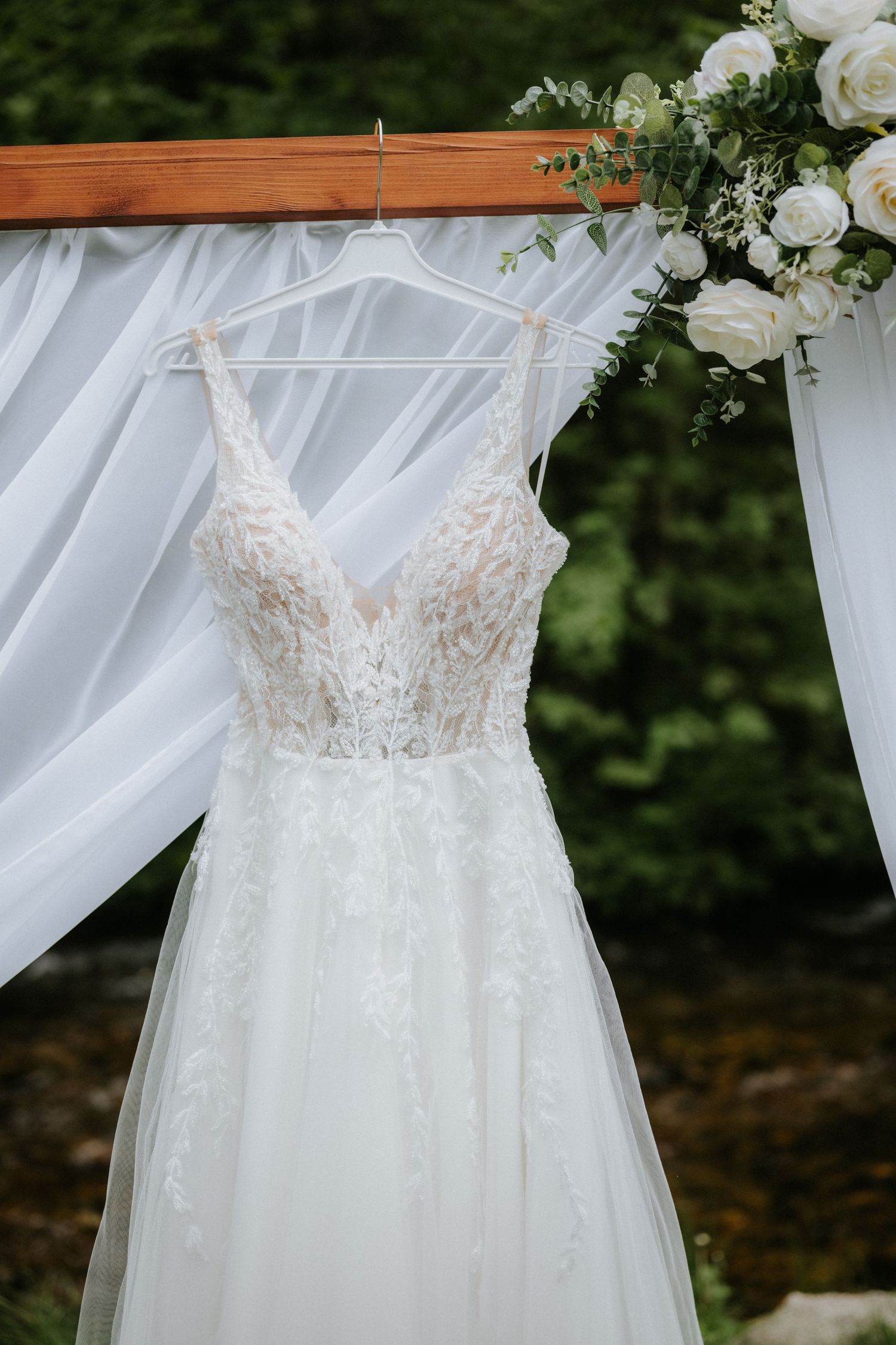 Elegant lace A-line wedding dress hanging on a wooden arch decorated with white roses and flowing drapes.