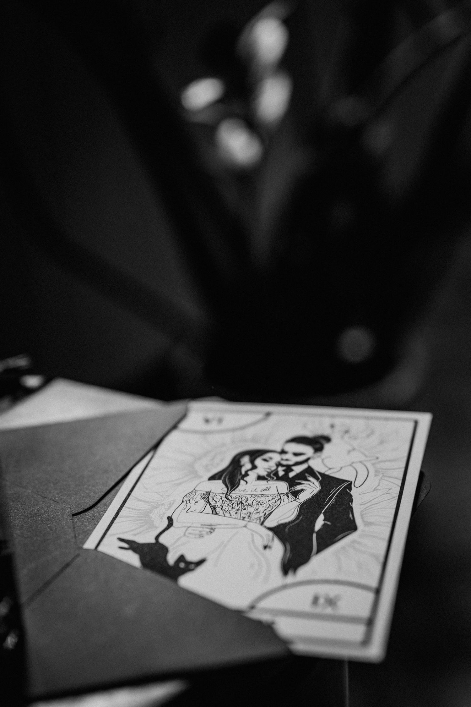 Black and white photo of a illustrated wedding card featuring an embracing couple, placed on a dark envelope.