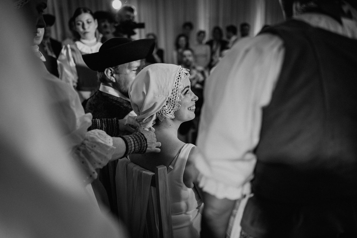 Black and white wedding photo of bride in headpiece surrounded by guests during traditional ceremony celebration.