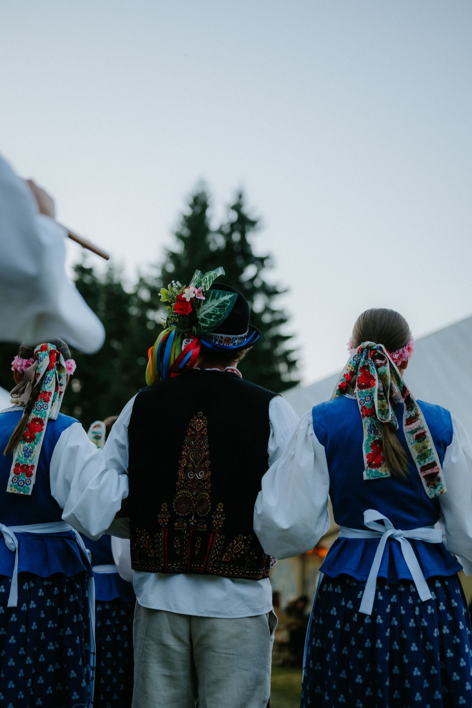 Three people in traditional Polish folk costumes with floral headpieces seen from behind at an outdoor cultural event.