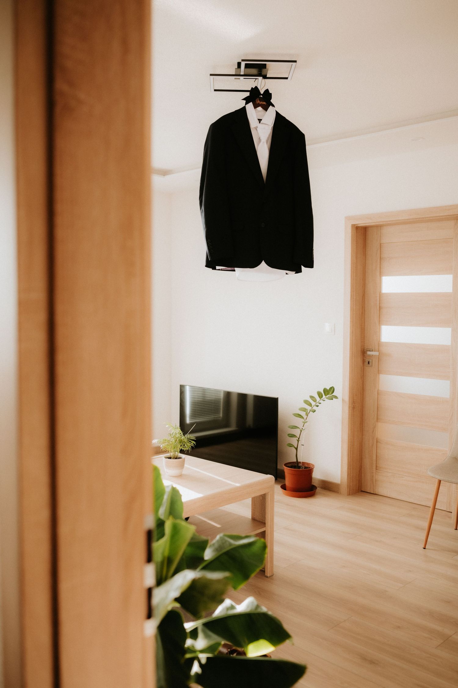 Black tuxedo jacket hanging on ceiling mount in modern living room with wooden furniture and indoor plants.