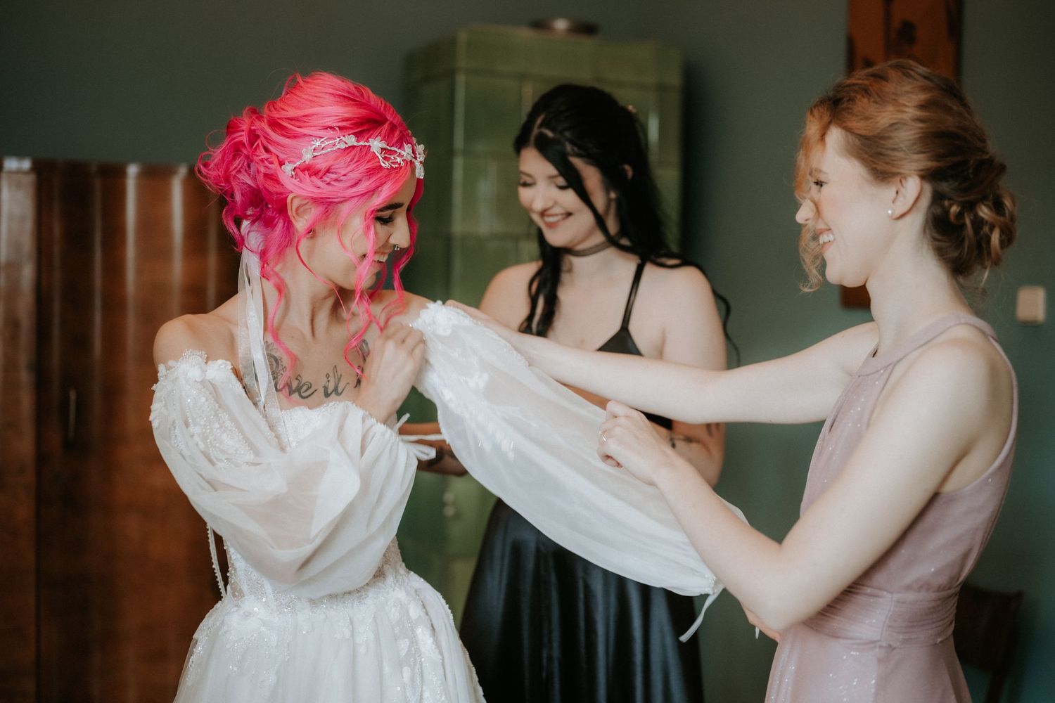 Bride with pink hair being helped into white wedding dress by two bridesmaids in black and pink gowns.