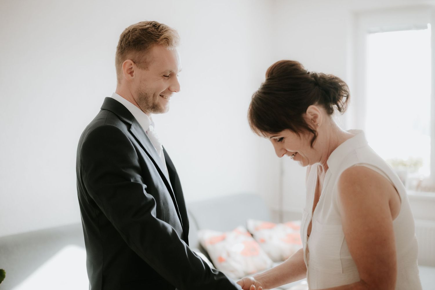 Bride and groom holding hands and smiling during an intimate wedding ceremony in a bright white room.
