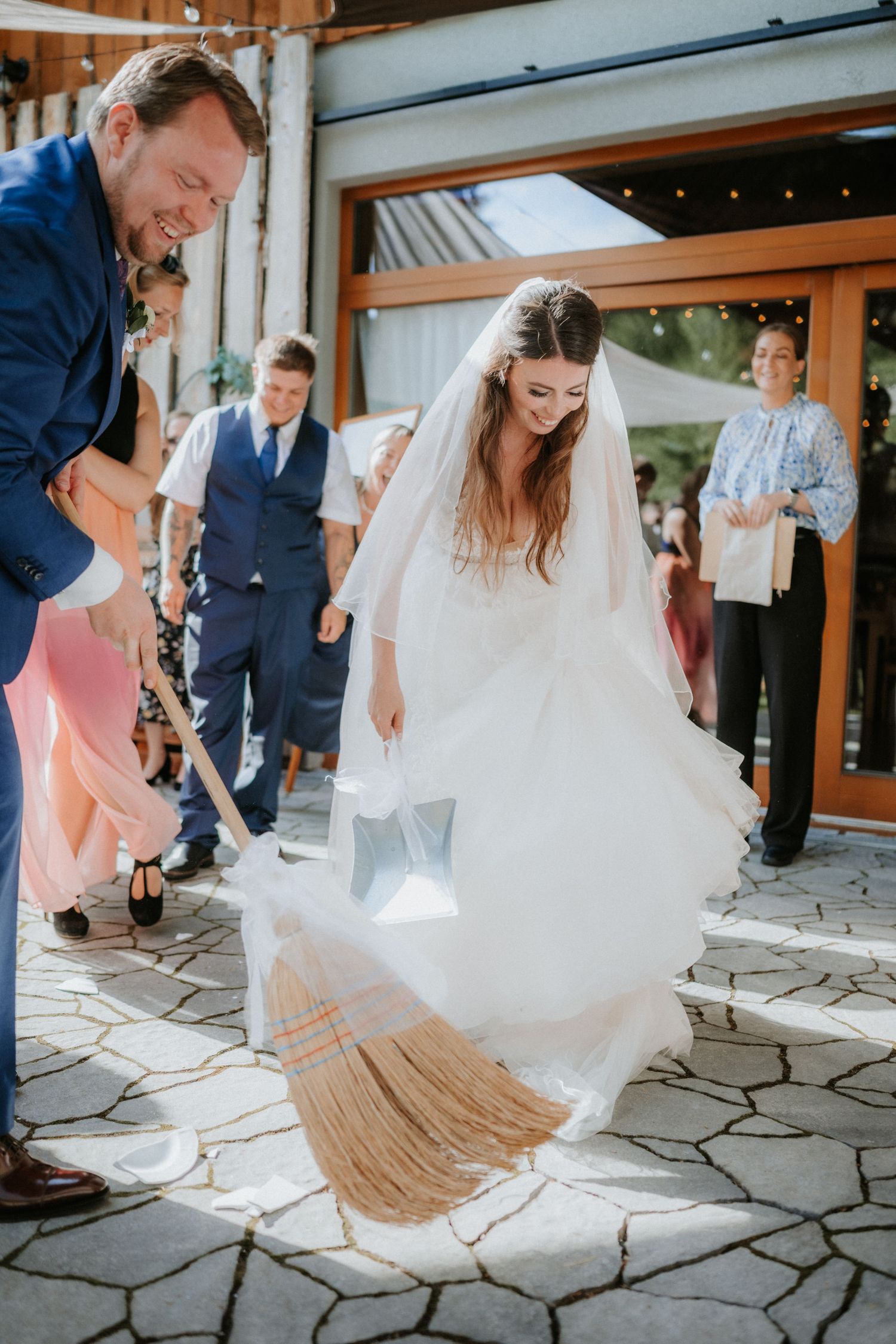 Bride and groom jumping over a broom in a traditional wedding ceremony outdoors, surrounded by smiling guests.