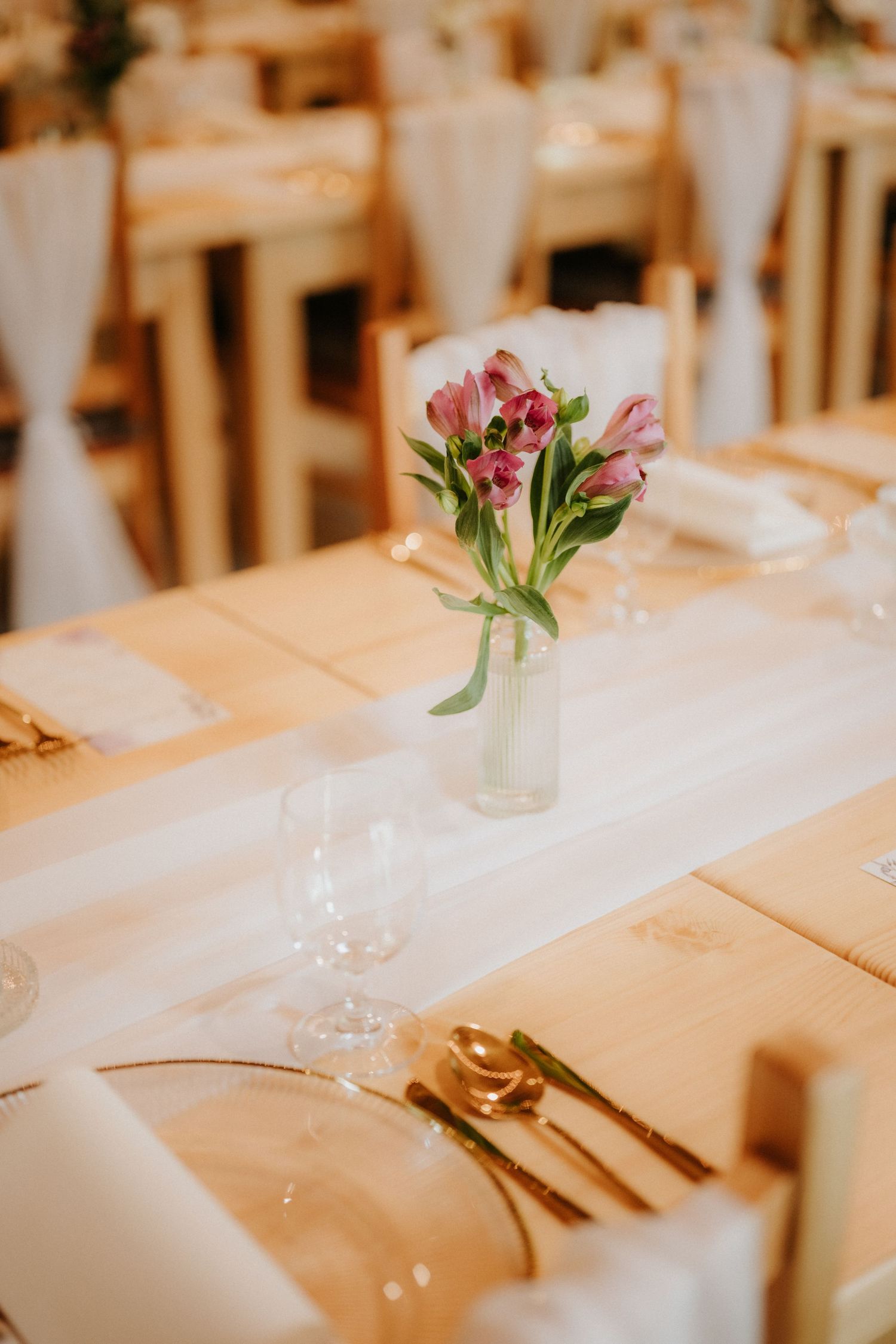 Elegant wedding reception table set with gold cutlery, crystal glasses, white linens, and a small vase of pink tulips.
