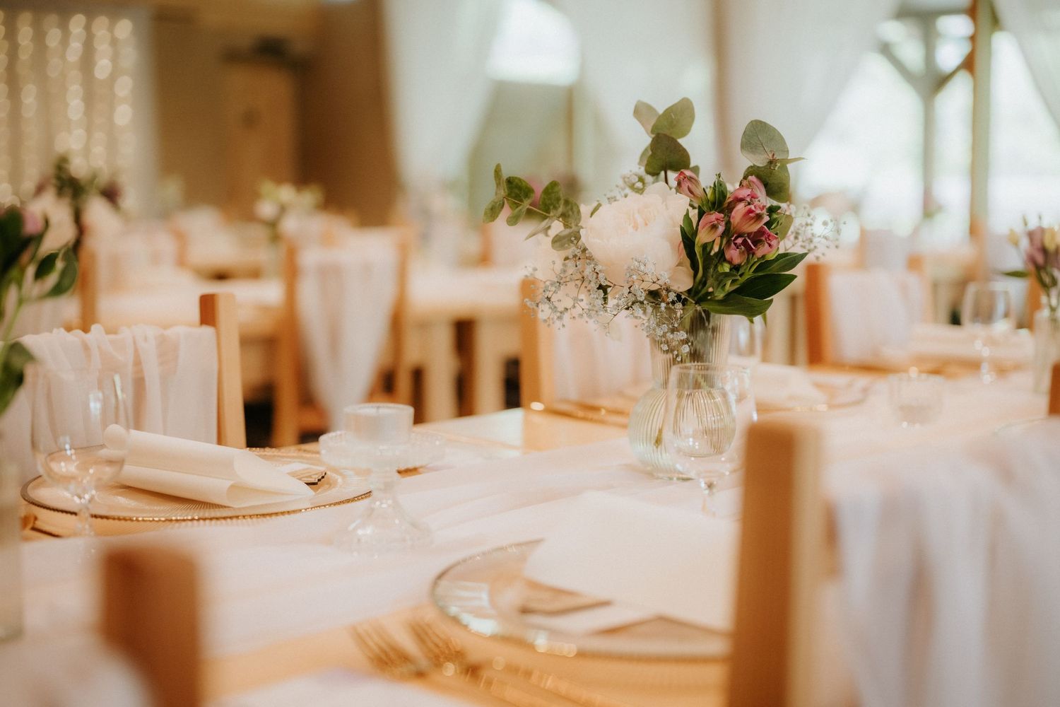 Elegant wedding reception table set with white linens, gold accents, and a floral centerpiece of pink roses and greenery.