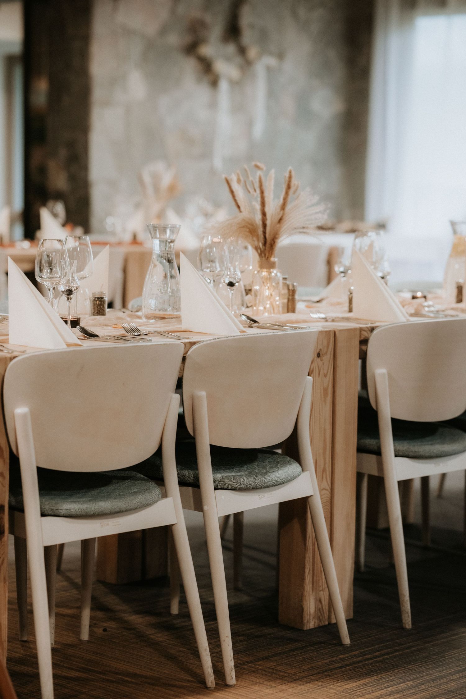 Elegant wedding reception table set with white chairs, glassware, and pampas grass centerpieces in a neutral-toned venue.