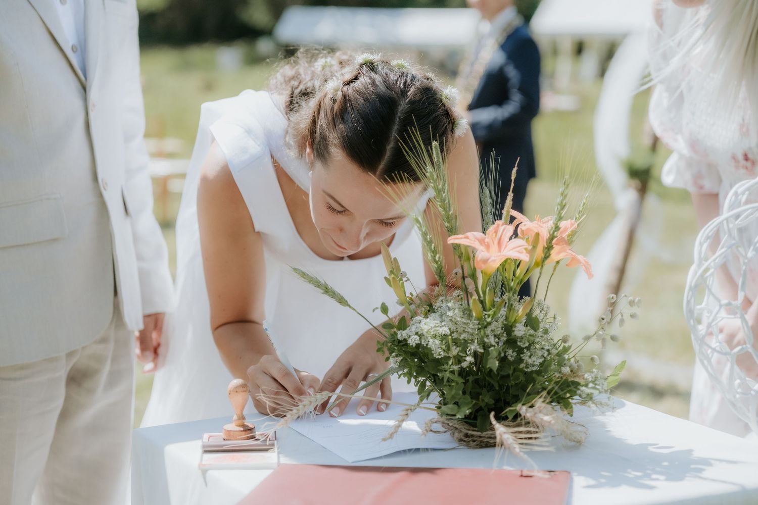 Bride in white dress signs wedding register at decorated outdoor table with peach floral arrangement.