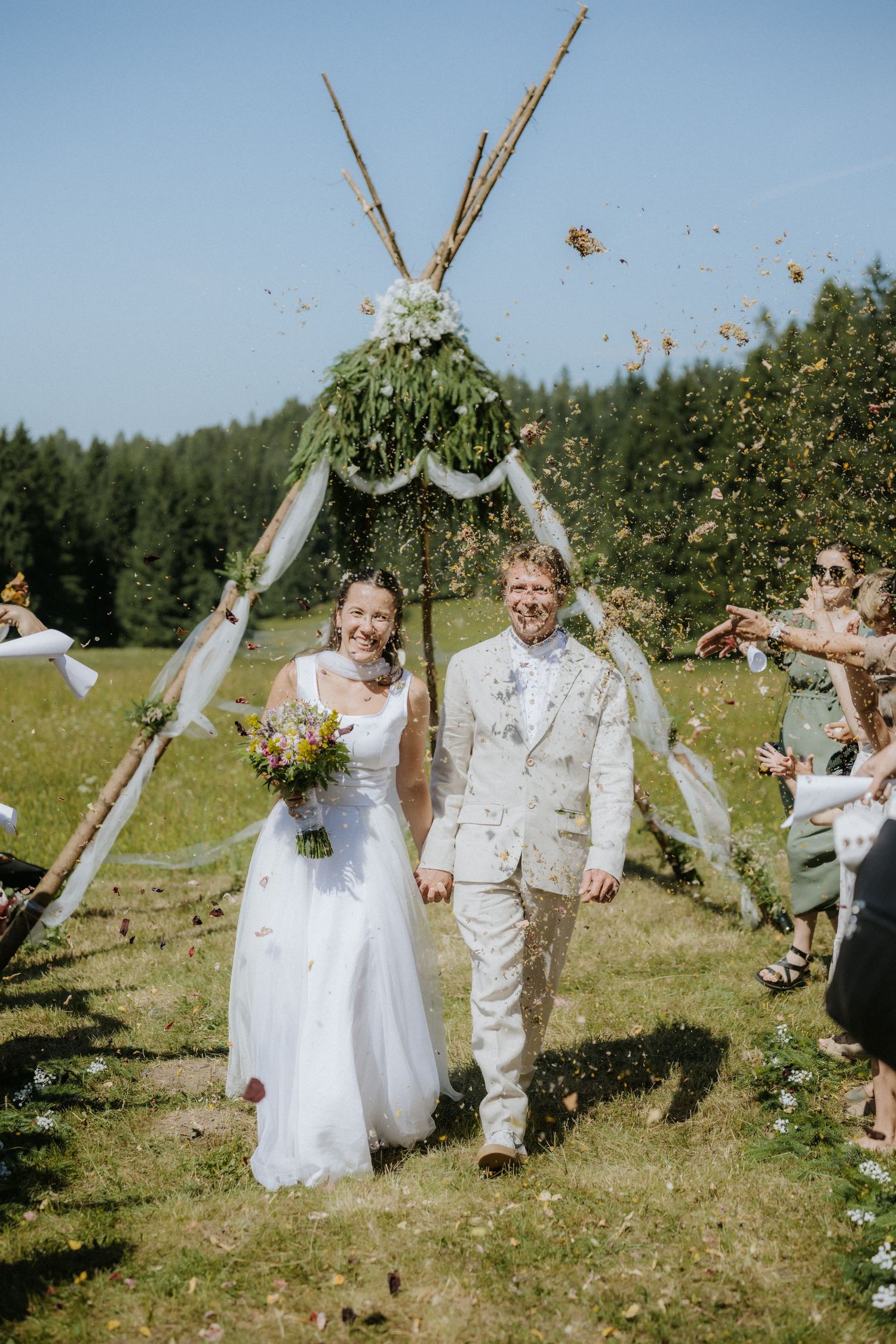 Bride and groom walk through confetti shower after outdoor bohemian wedding ceremony with teepee arch.