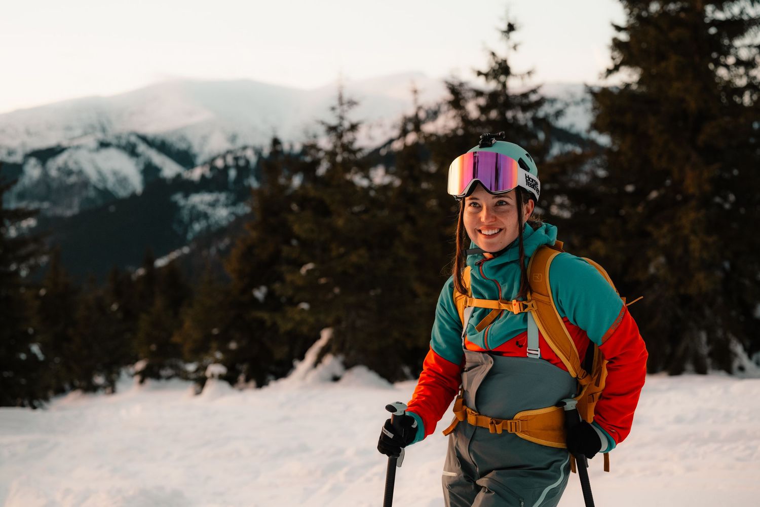 Smiling skier wearing colorful jacket, pink goggles, and helmet with backpack in snowy mountain landscape.