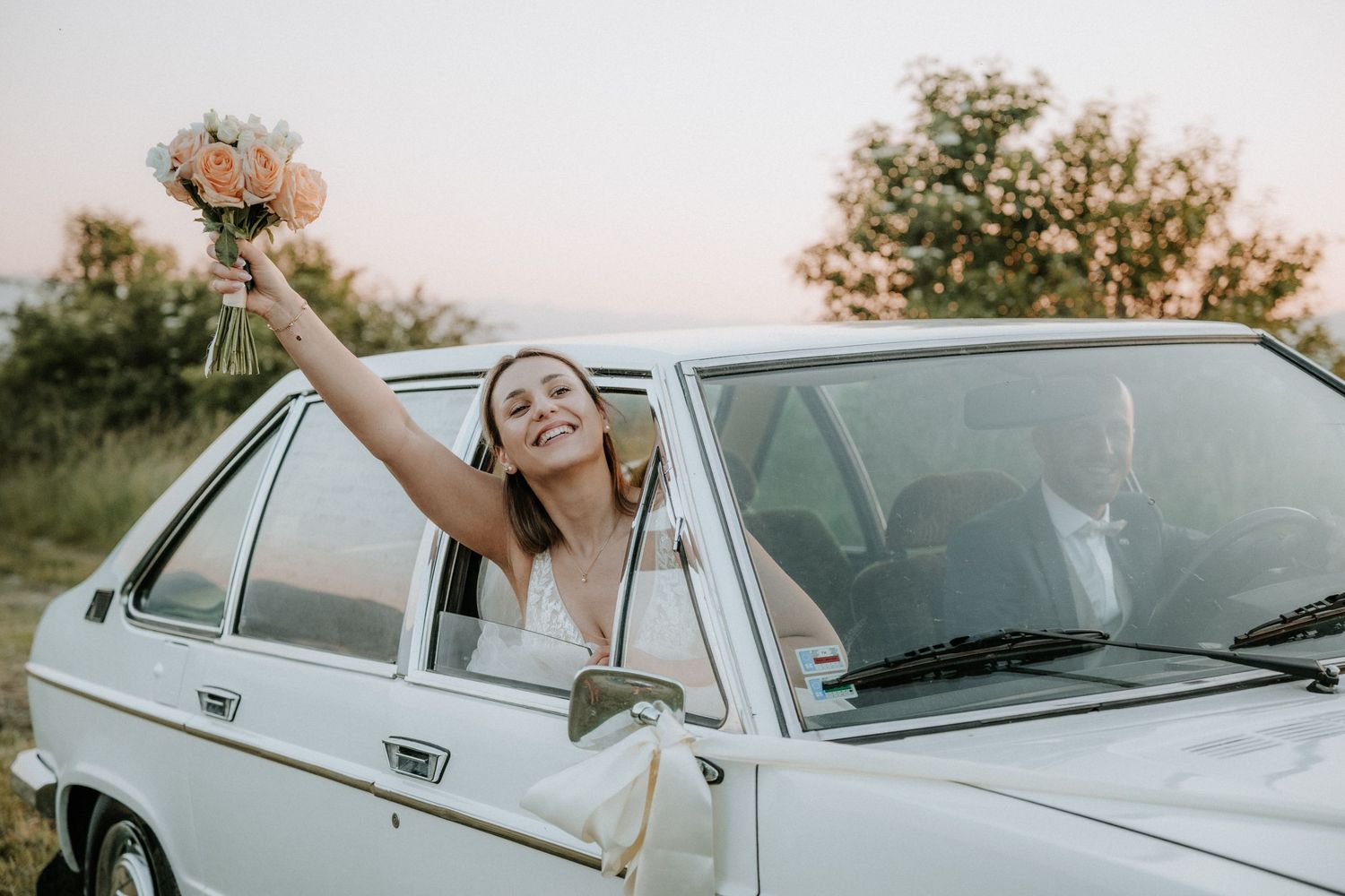 Joyful bride leaning out of a white vintage car window, holding a bouquet aloft at sunset on her wedding day.