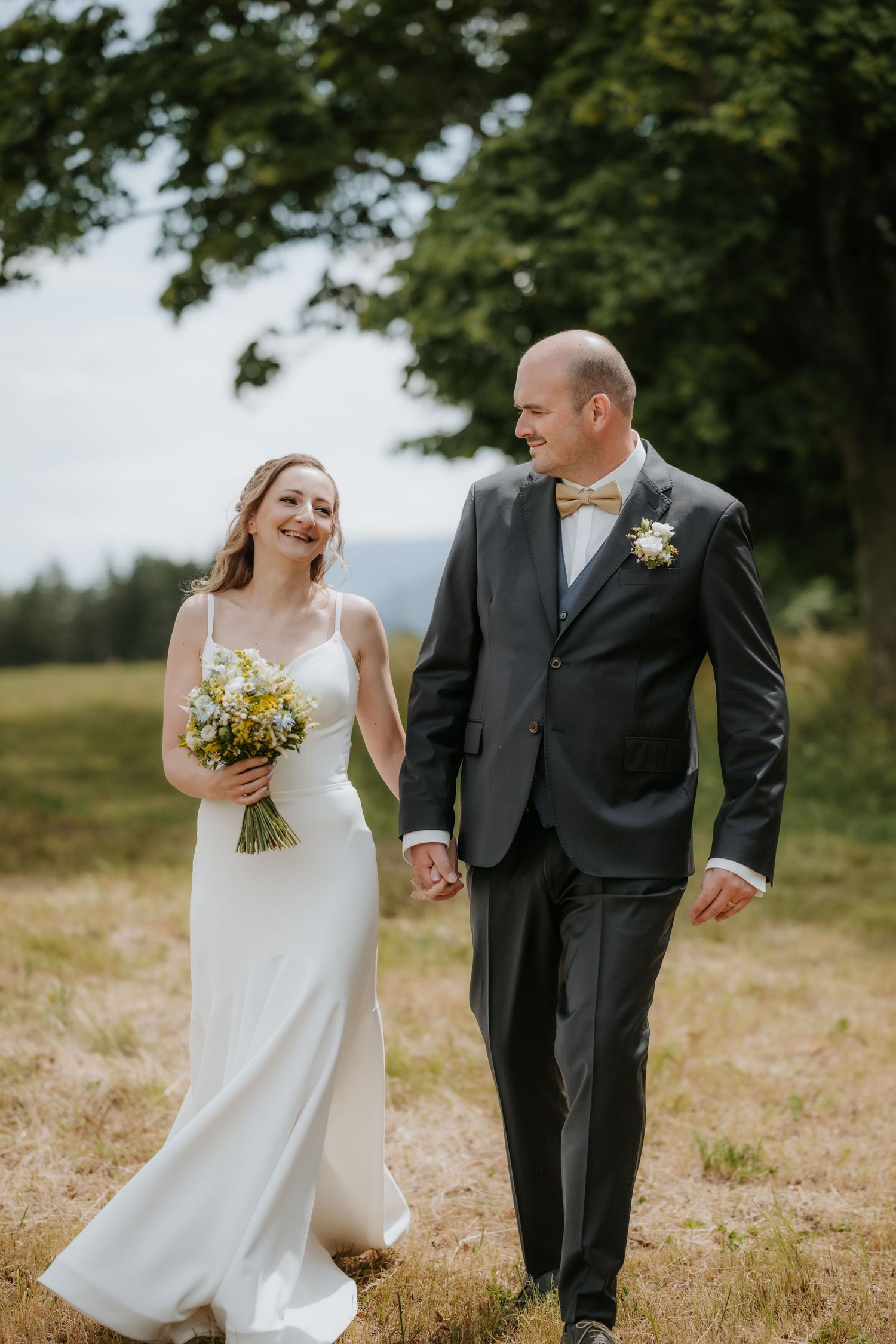 Bride and groom walk hand-in-hand through a sunny meadow, bride holding yellow wildflower bouquet in white gown.