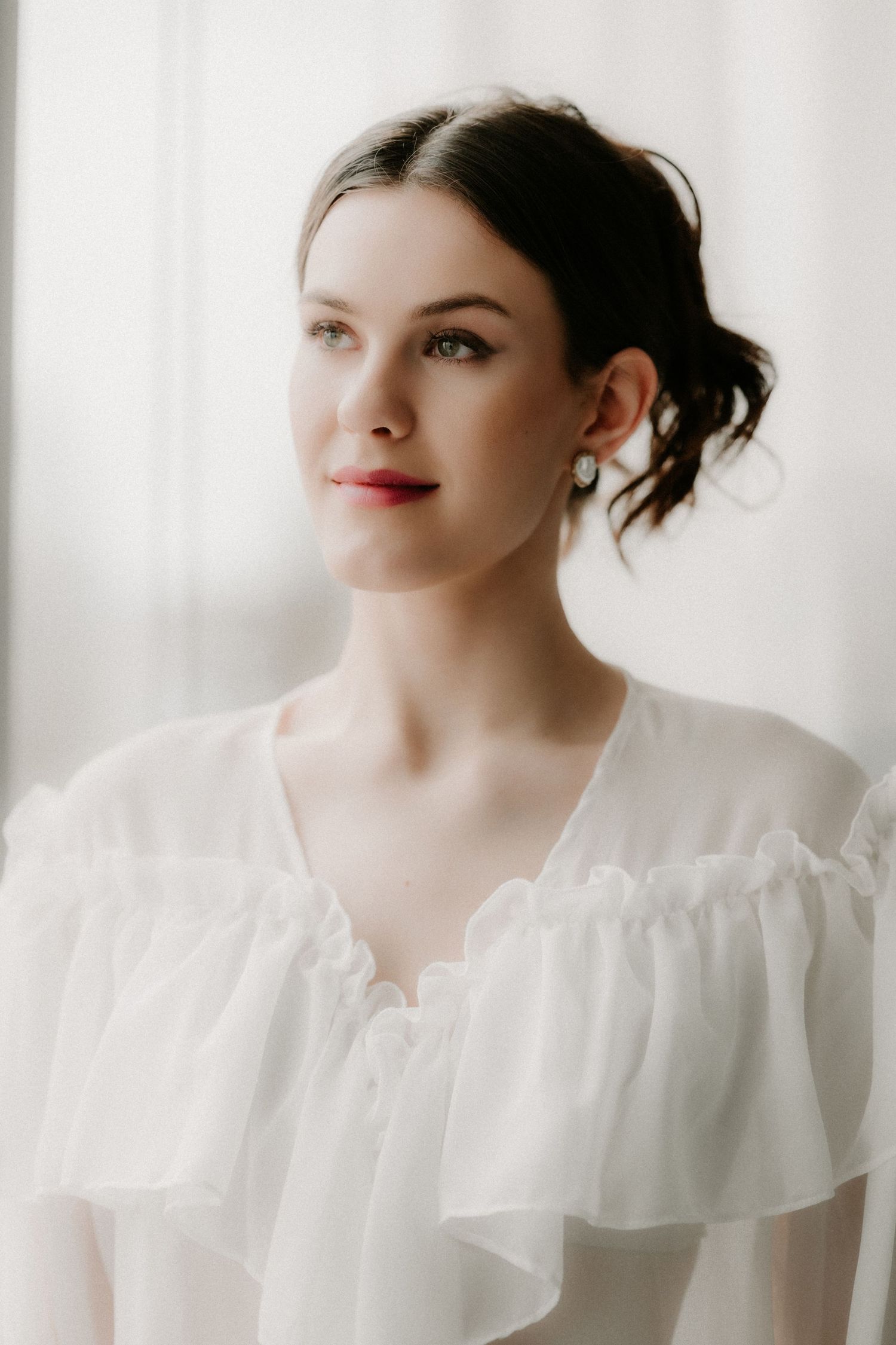 Elegant young woman in a white ruffled chiffon blouse with an updo hairstyle, gazing softly to the side near a window.