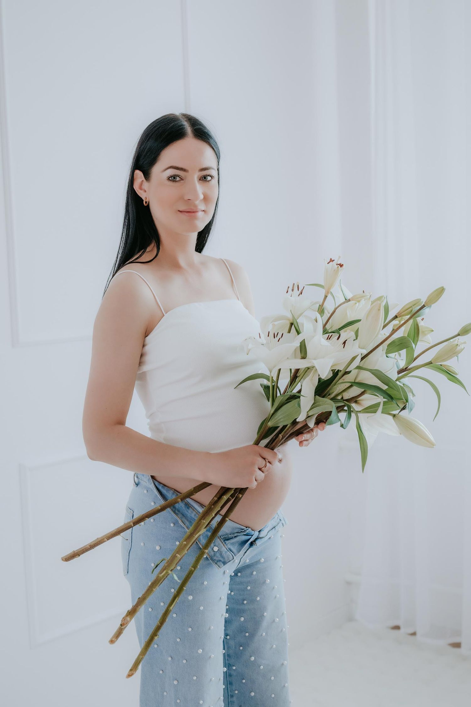 Pregnant woman holding white lily bouquet in white crop top and jeans.