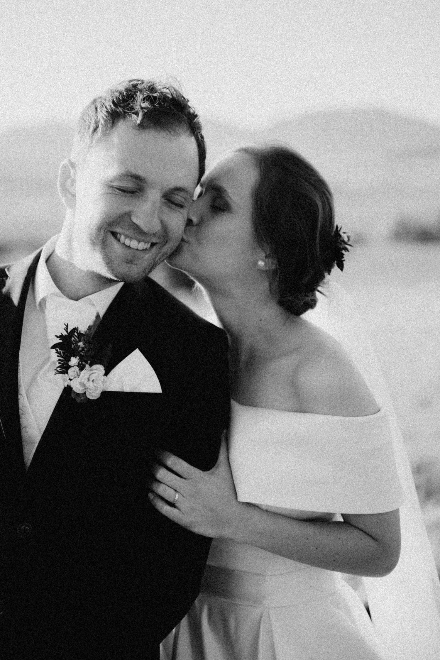Black and white wedding photo of bride kissing groom on cheek while he smiles joyfully.