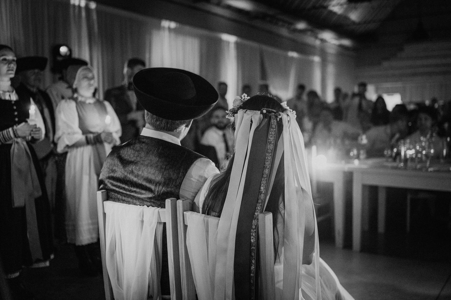 Black and white photo of a couple in traditional folk wedding attire during a ceremony with guests in background.