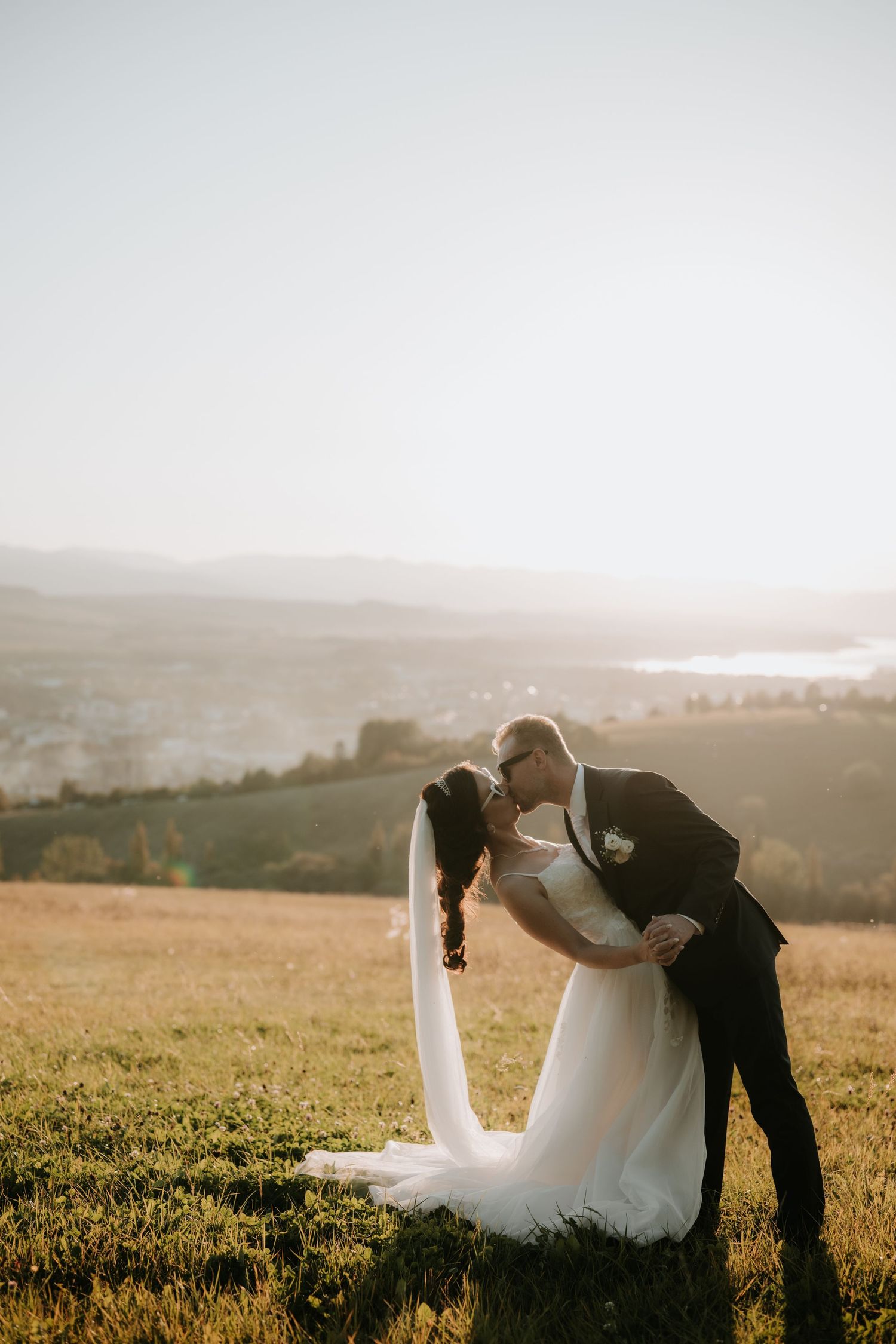 Bride and groom share a romantic dip kiss on a sunlit hillside with a scenic valley and mountains in the background.