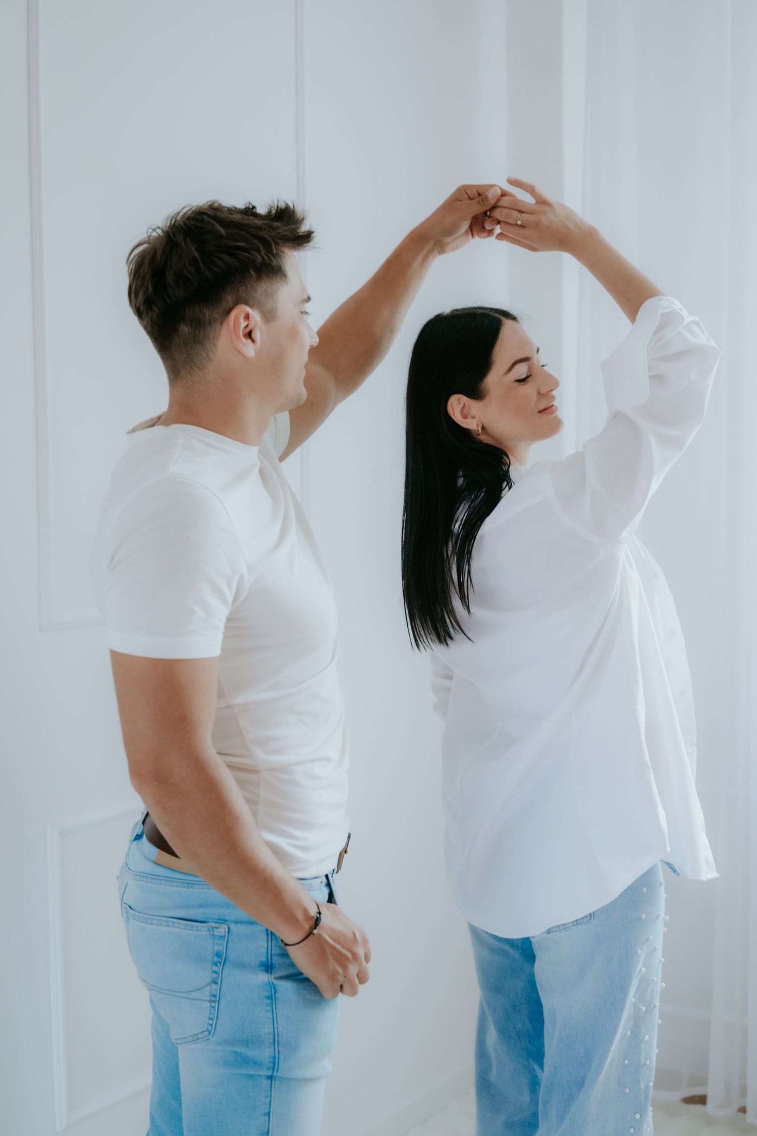 A couple in white tops and jeans dancing together in a bright white room.