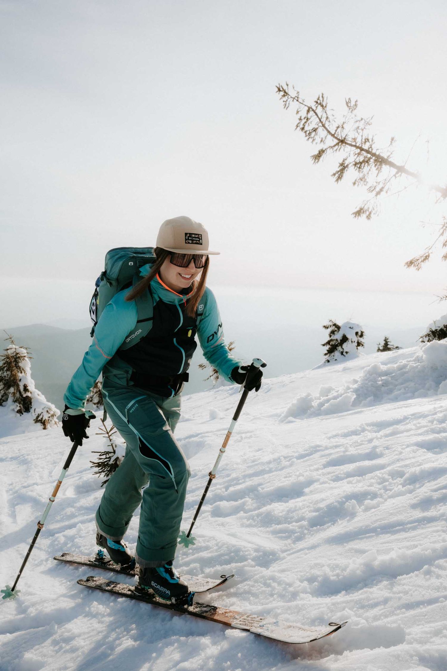 Woman in teal ski gear and backpack skiing uphill on a snowy mountain slope on a bright winter day.