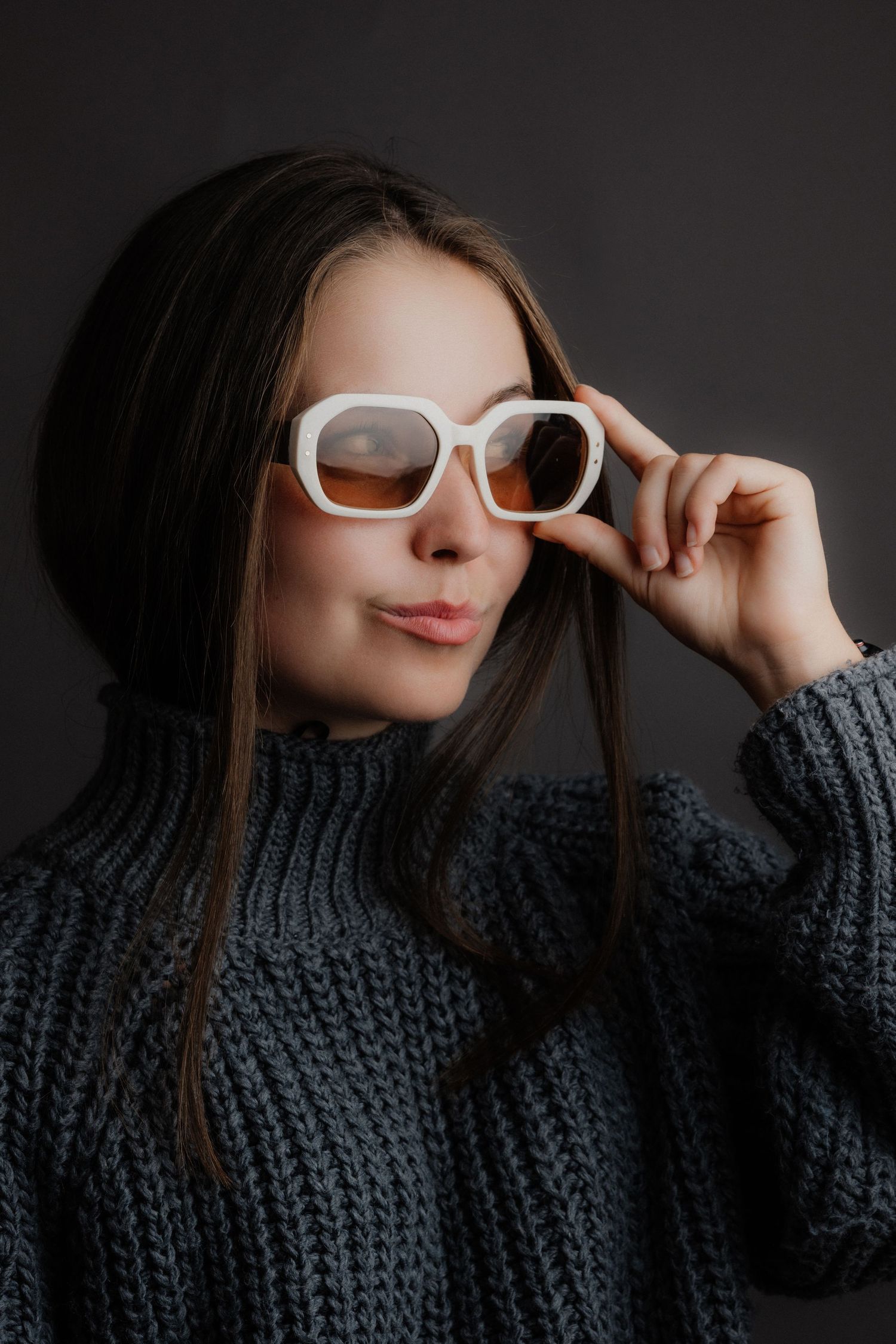 Young woman in gray knit sweater adjusting white square sunglasses with orange tinted lenses against dark background.