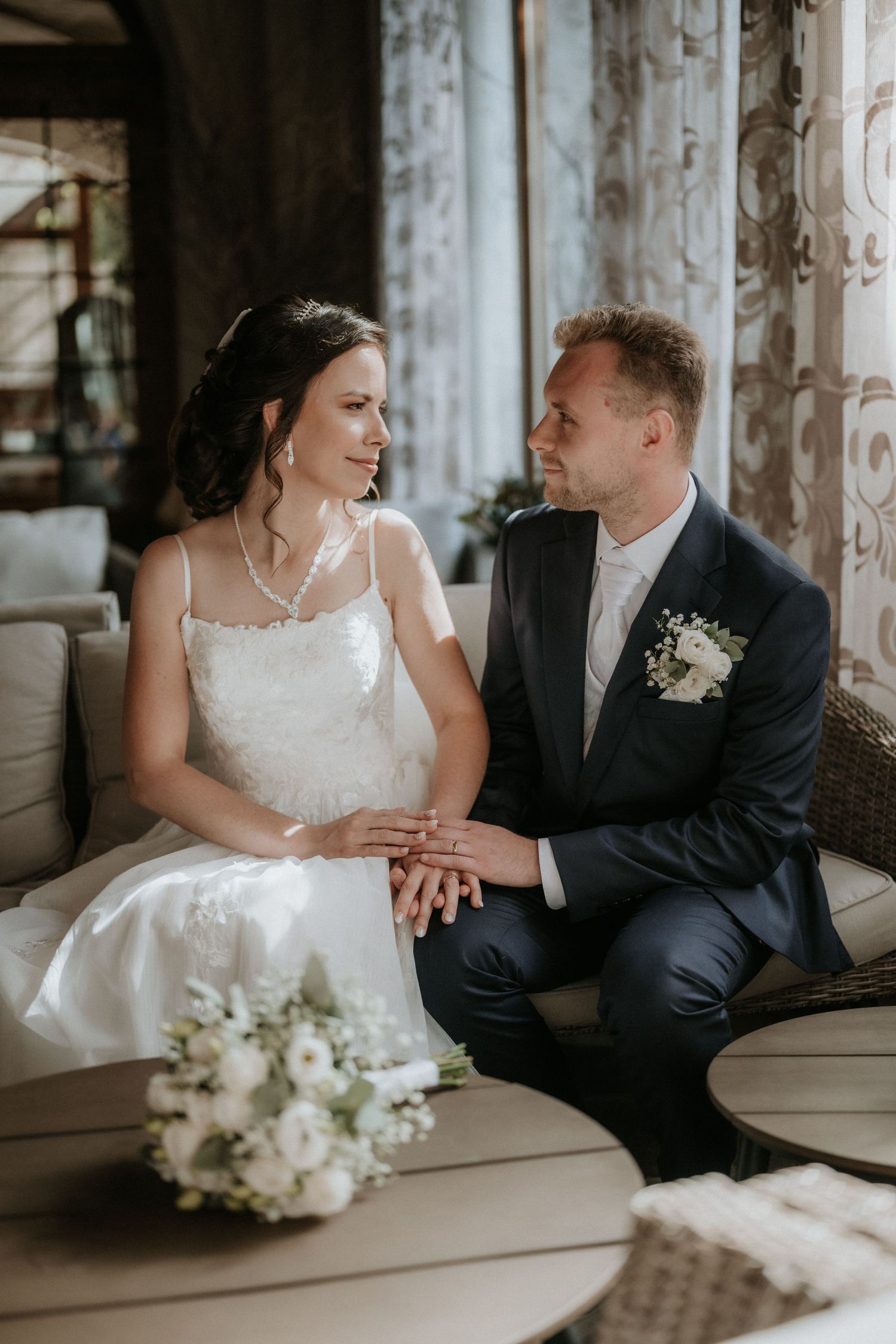 Bride and groom gazing at each other while seated on a sofa with bouquet in the foreground.