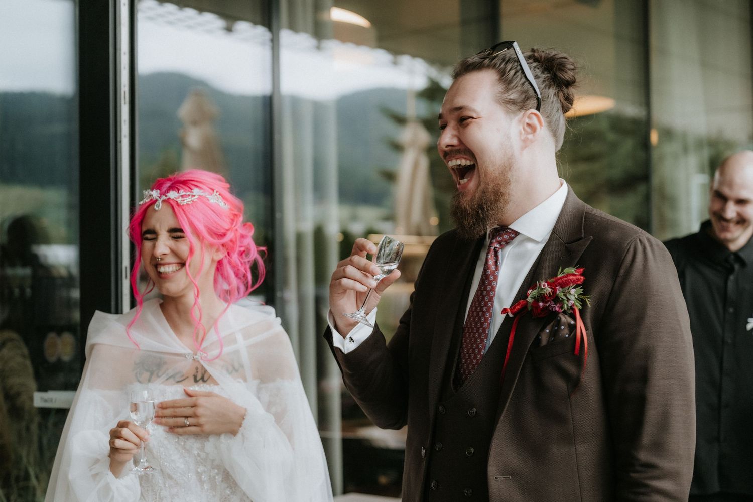 Laughing bride with pink hair and groom in brown suit with red boutonniere celebrating at wedding reception.